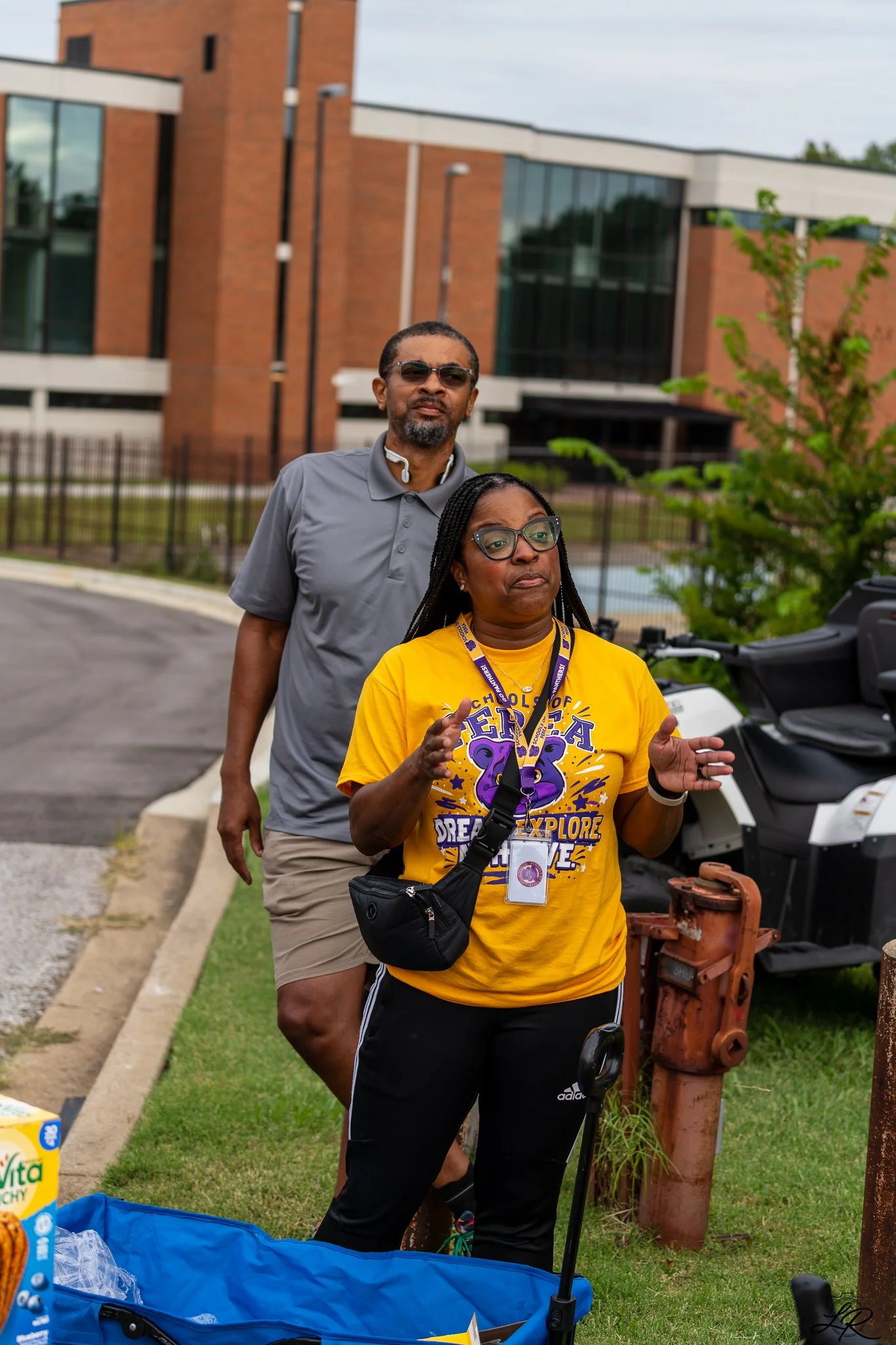 Two women standing outdoors near a sidewalk, with a modern brick and glass building in the background. One woman is wearing sunglasses and a gray shirt, and the other woman is wearing glasses, a yellow T-shirt, and has a camera around her neck. There