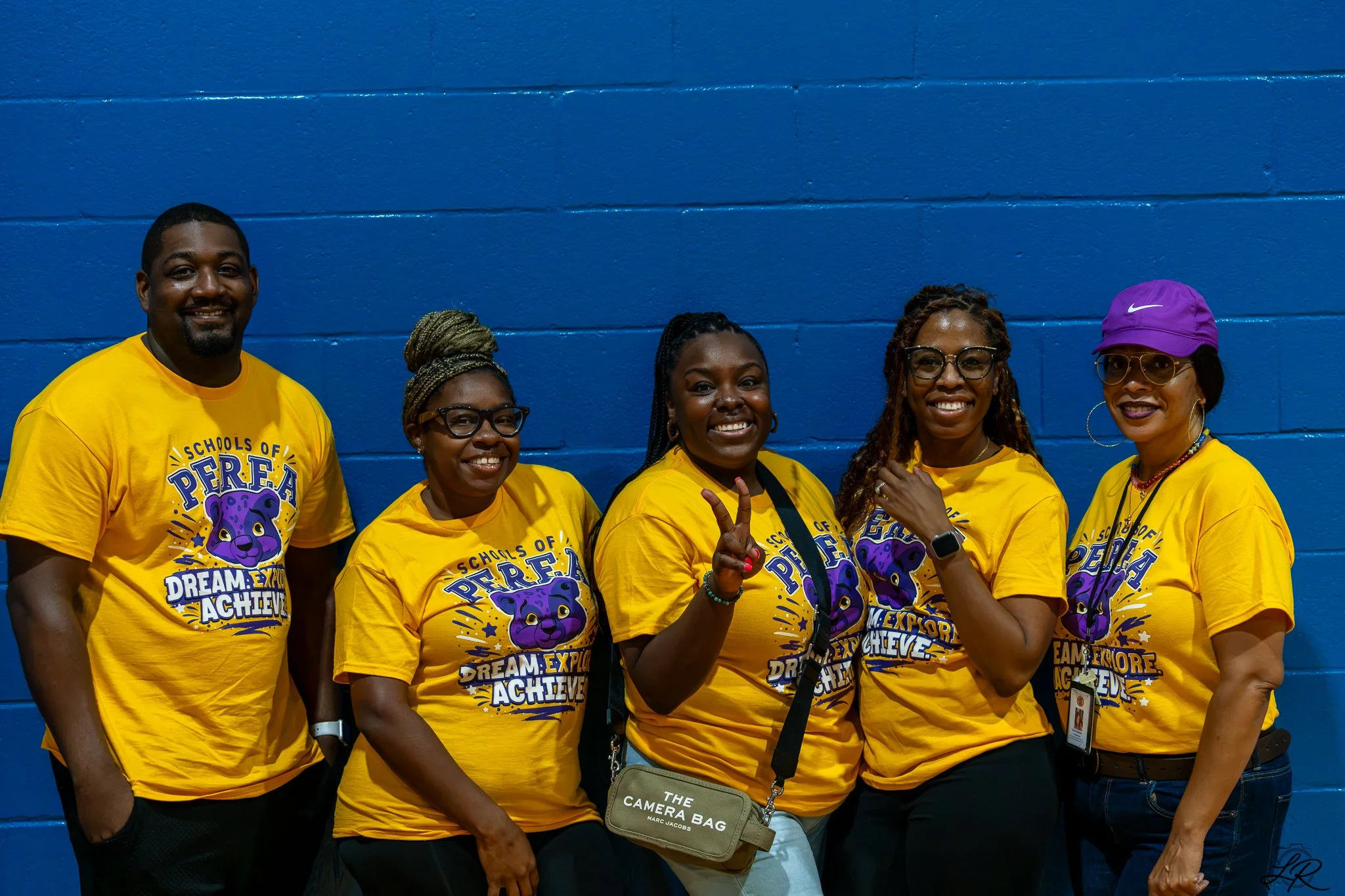Group of six diverse people standing against a blue wall, wearing matching yellow shirts with a purple lion logo and the text 'Schools of P.E.R.S.E.V.E.R.E. Dream Explore Achieve.' They are smiling, with some making peace signs, and one woman wearing