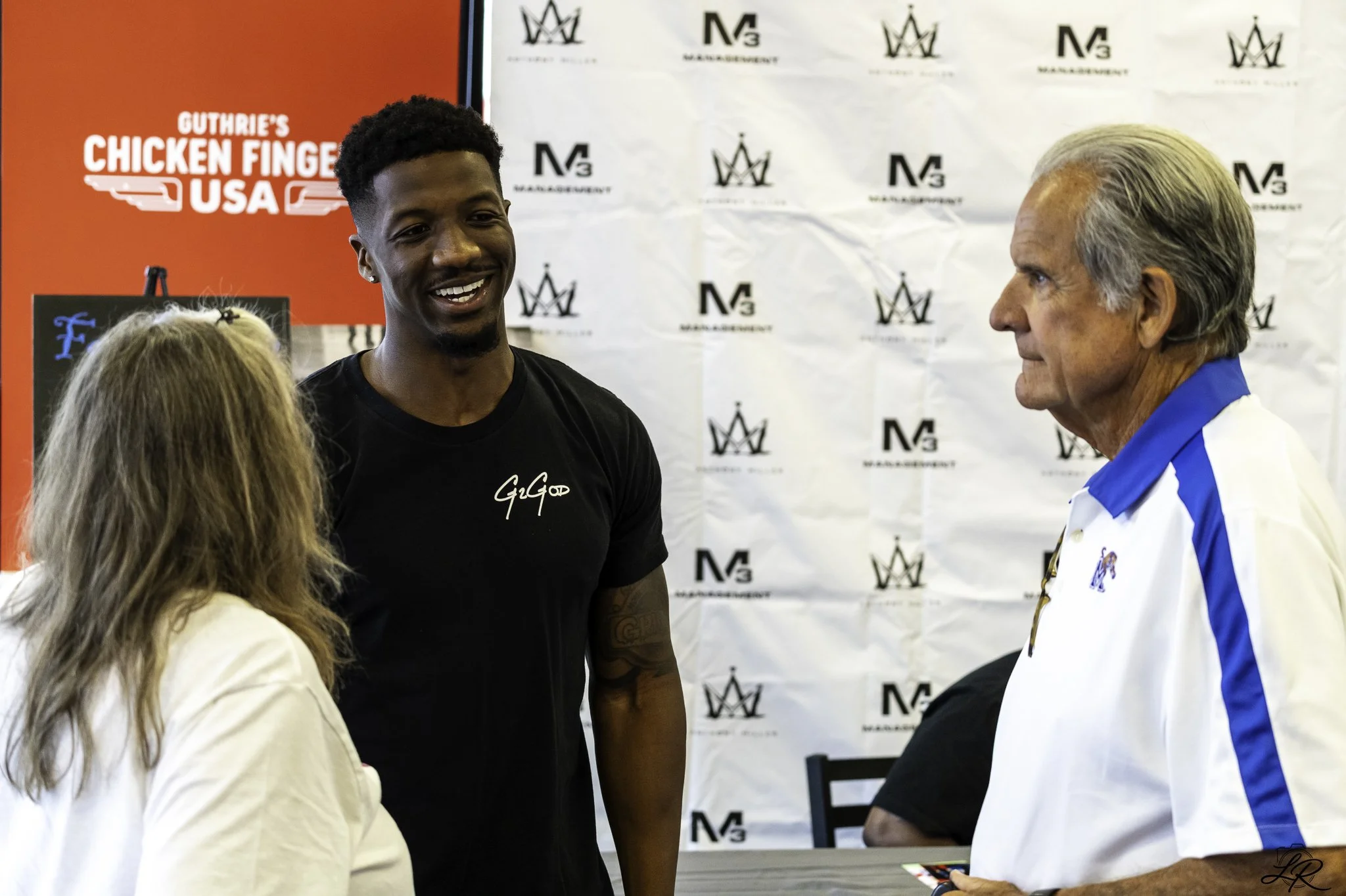 Three people are engaged in conversation at an event with a backdrop featuring logos. One woman with blonde hair is facing away from the camera, two men are facing her. One man has dark skin, short black hair, and is smiling, wearing a black shirt. T