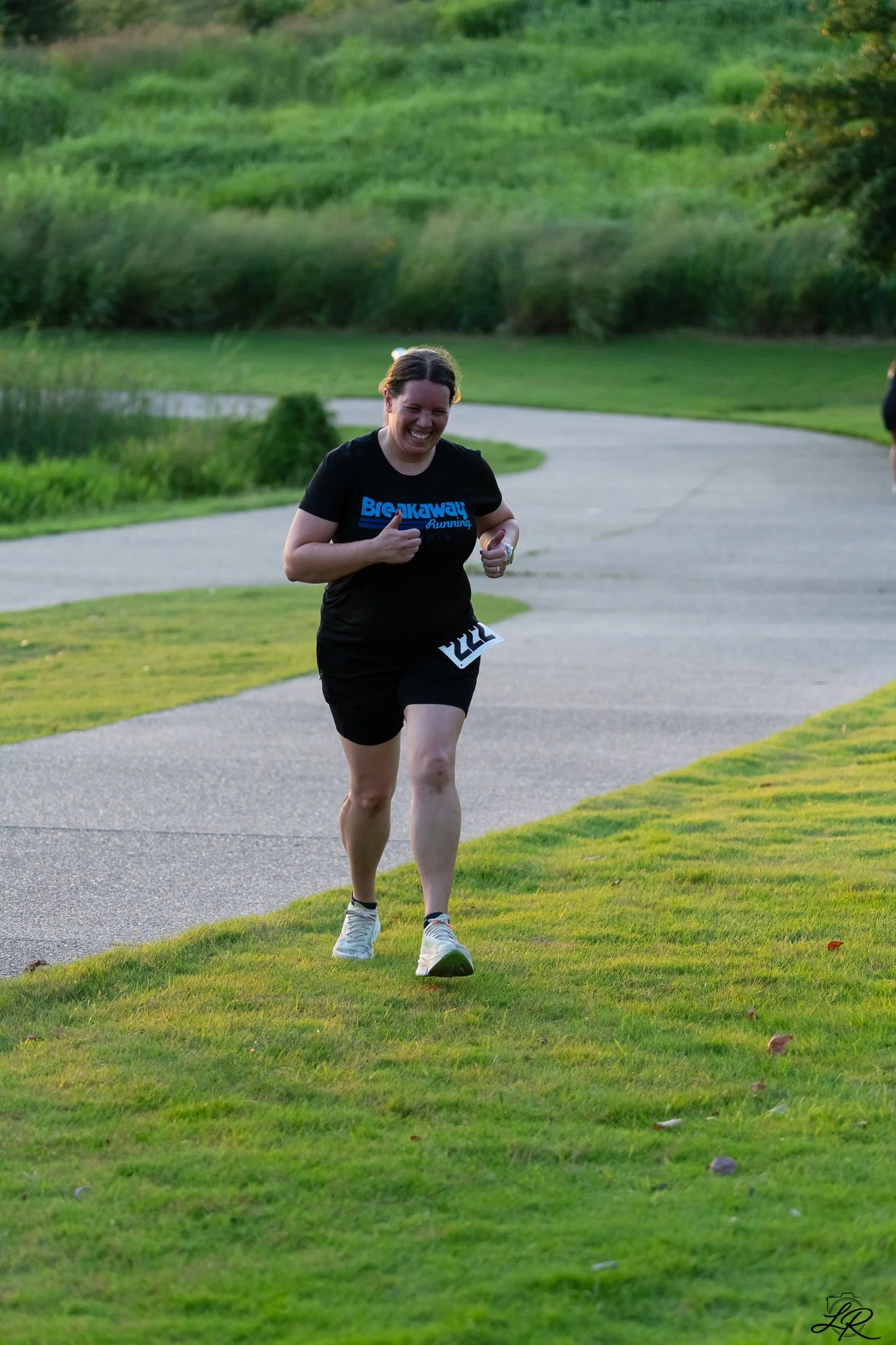 Woman running on a paved trail in a park with green grass and trees in the background, smiles, wearing a black T-shirt and shorts.