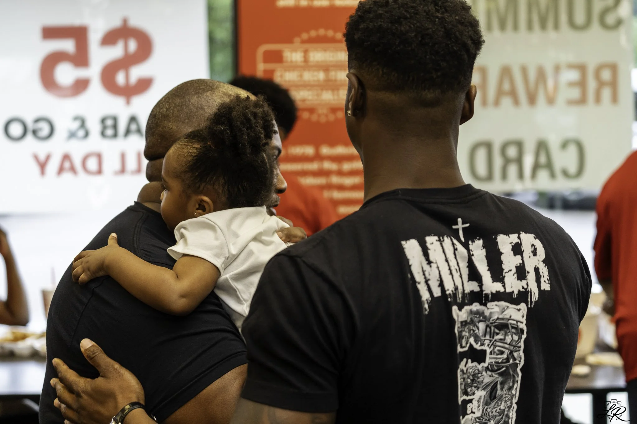 A person holding a child while another person stands nearby, all indoors with banners and signs in the background.