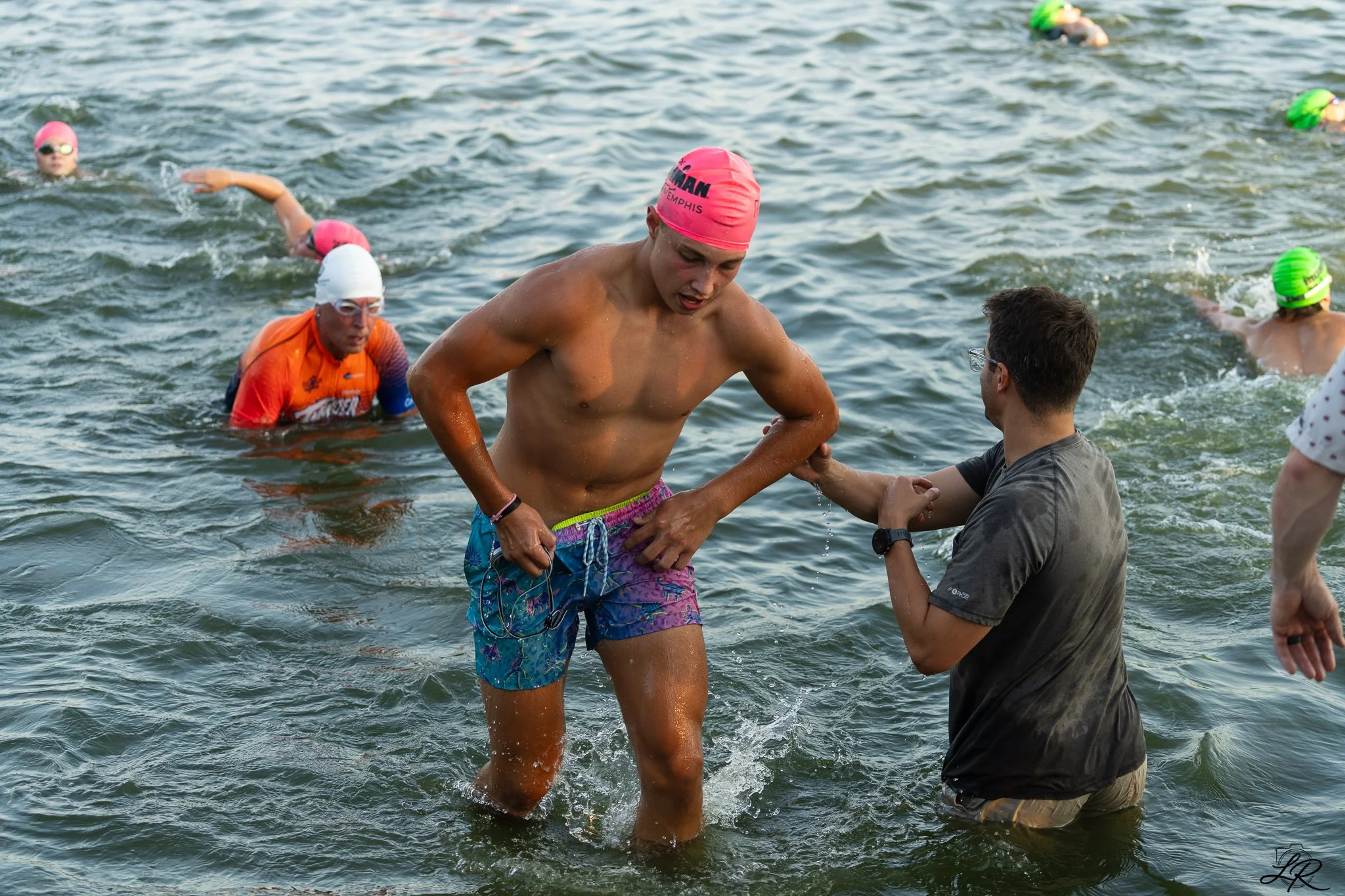 Swimmers in a body of water participating in a race or event, with one man in colorful swim trunks and a pink swim cap shaking hands with a man on the shore.