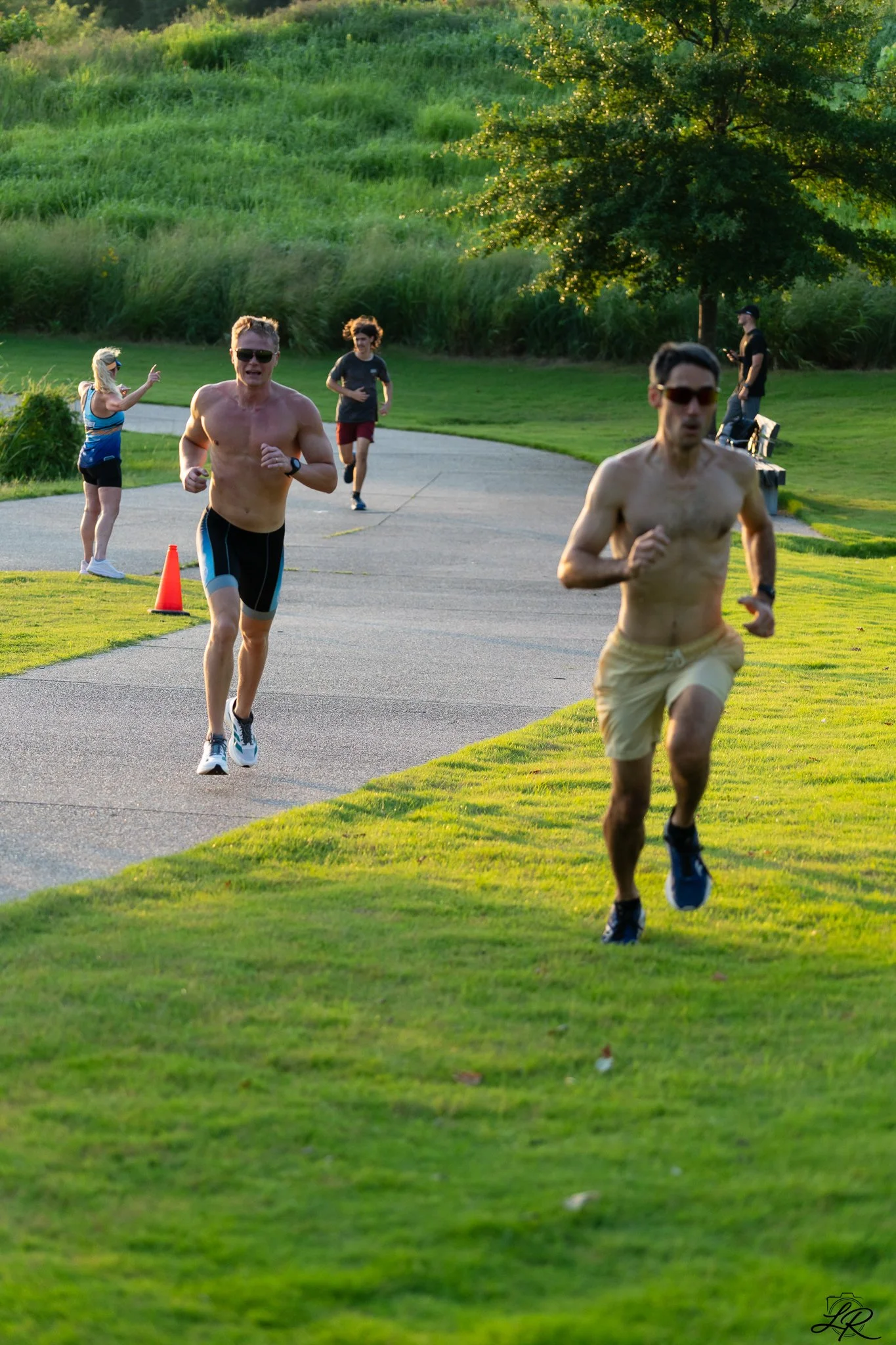 Two shirtless men running on a paved park trail during sunset. A woman in athletic wear and a man walking with a phone in the background. Green trees and grass surround the trail.