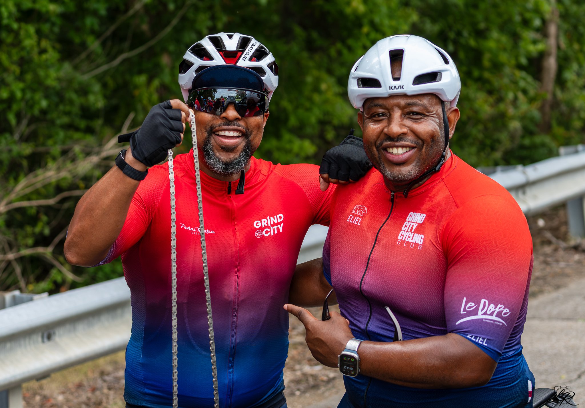 Two men wearing cycling helmets and matching red and purple shirts, smiling and posing outdoors with a background of green trees and a metal railing.