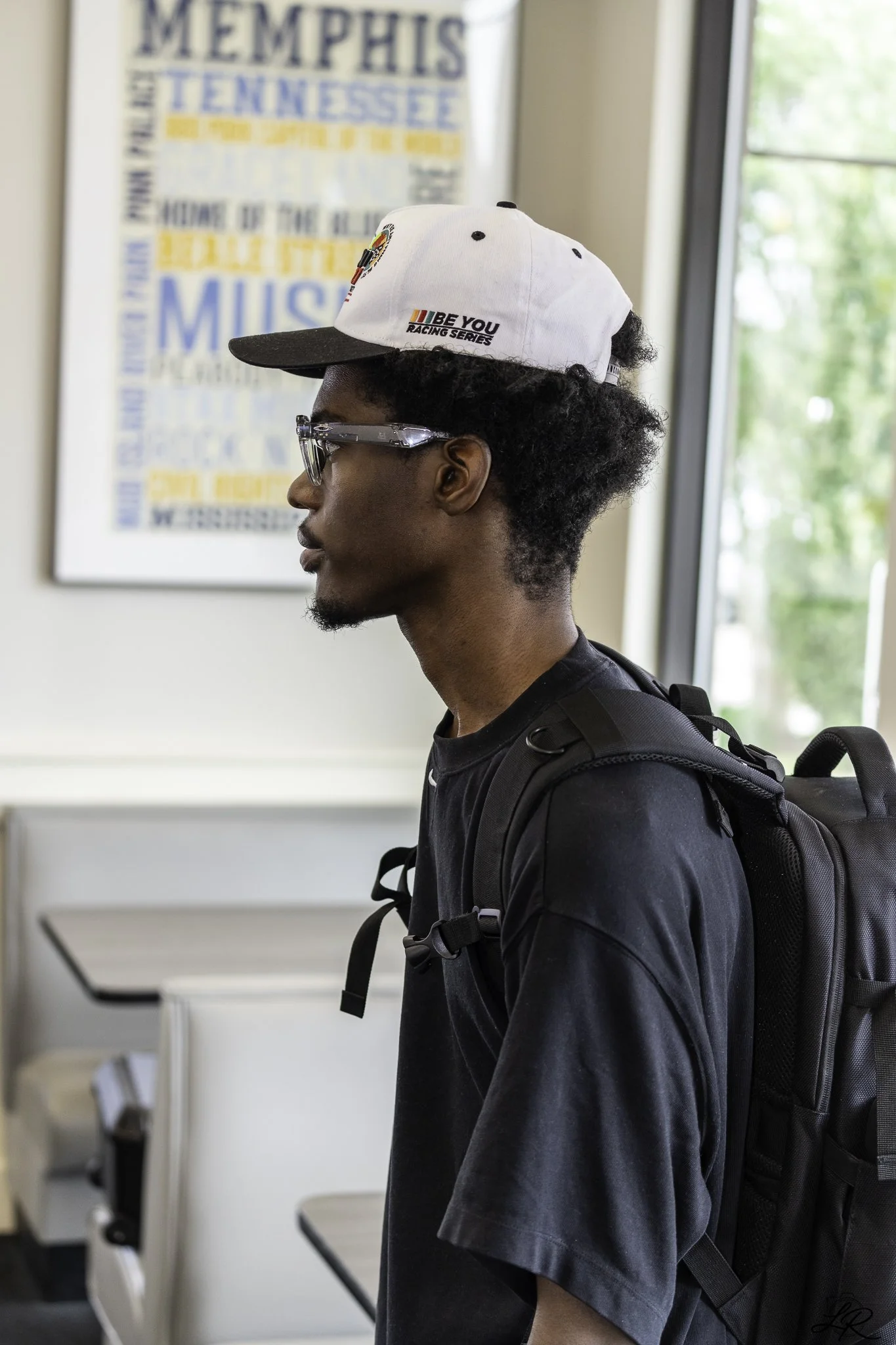 A young man with curly black hair, glasses, wearing a white cap with logos and text, a black t-shirt, and a backpack, standing indoors near a window with green trees outside, with a Memphis poster on the wall behind him.