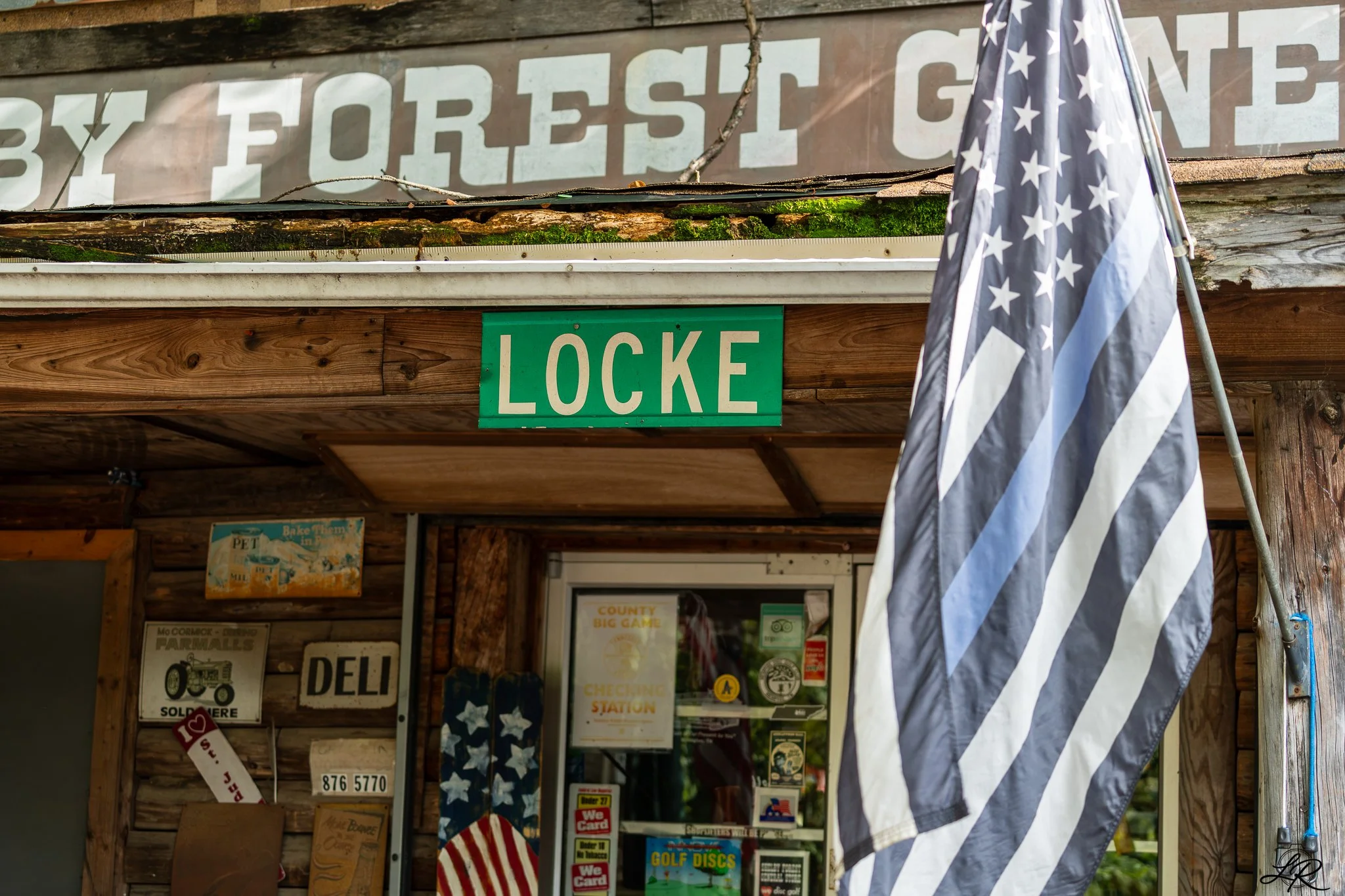 Wooden building corner with a green 'LOCKE' sign, posters in the window, an American flag, and a faded sign above.