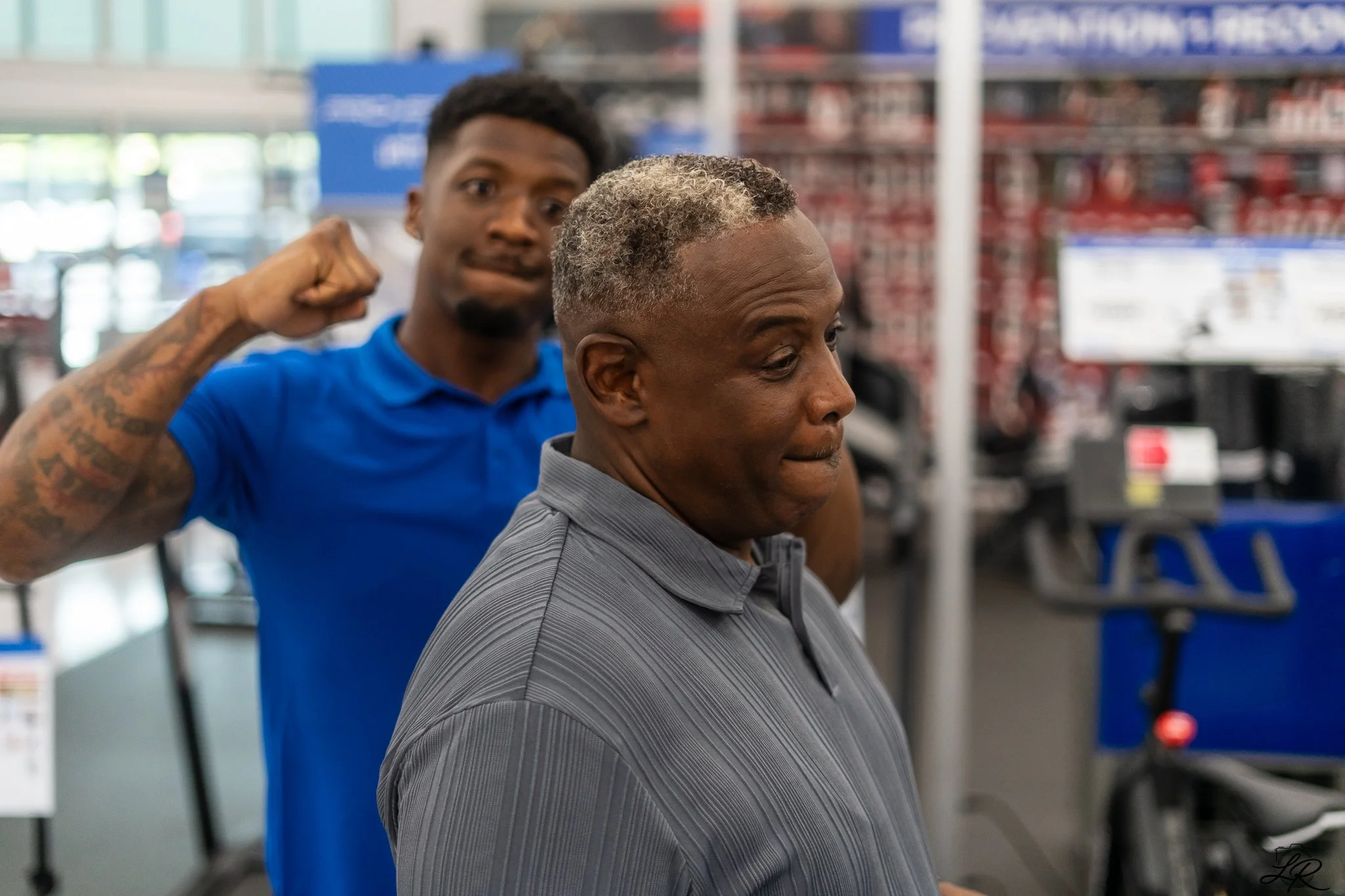 A man in a gray shirt gets a haircut at a store while a barber in a blue uniform poses behind him making a fist with his right hand. The background shows shelves with boxed products.