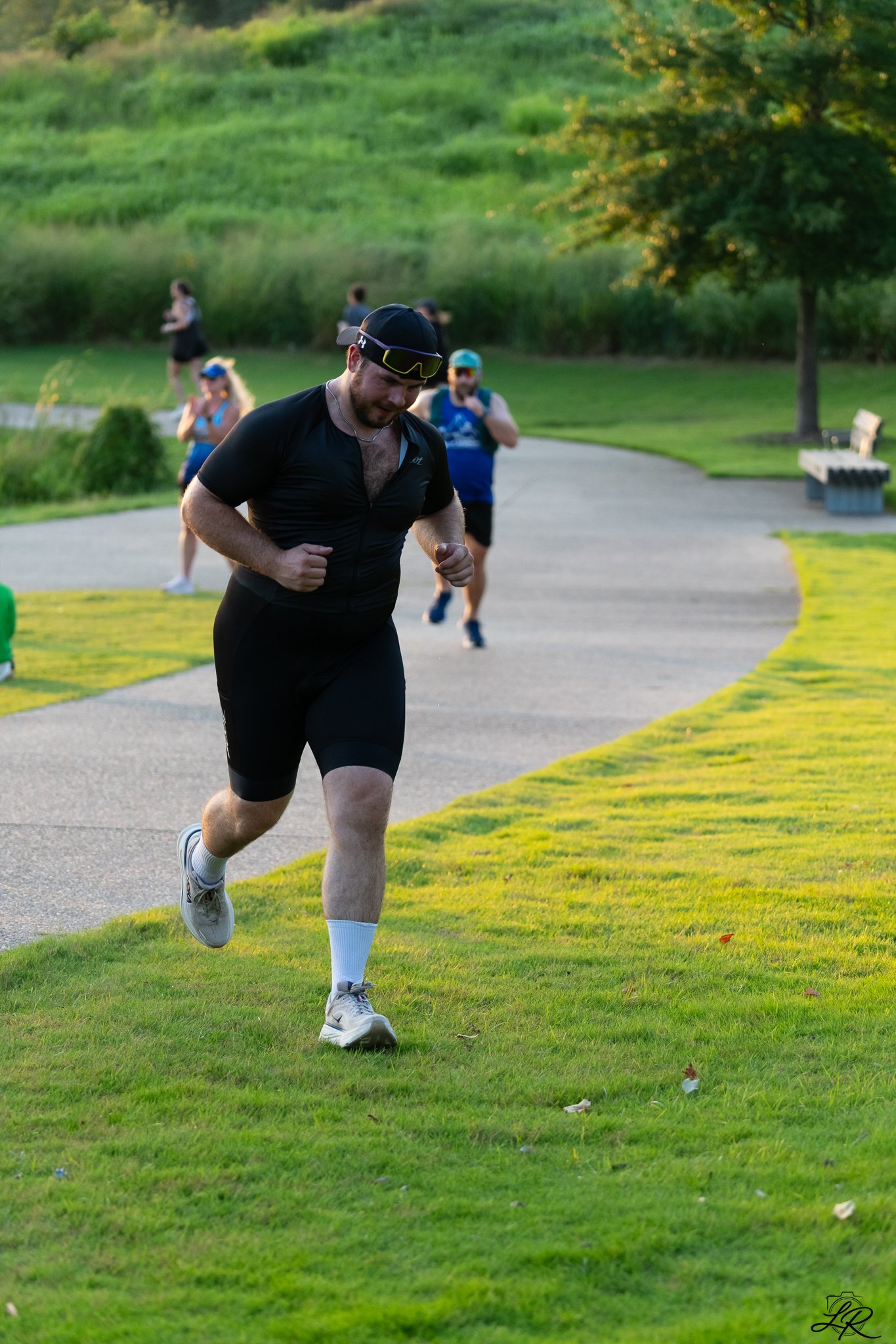 A man in a black athletic shirt and shorts runs on a park path during daytime, with a green landscape and trees in the background.