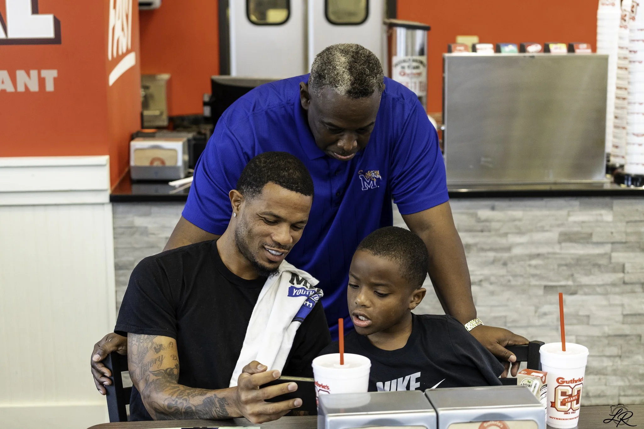 A man with a tattoo on his arm wearing a black Nike shirt, and an older man with gray hair wearing a blue shirt, are sitting at a table together, looking at a smartphone. A young boy in a black shirt is also looking at the phone. Two drinks with red 