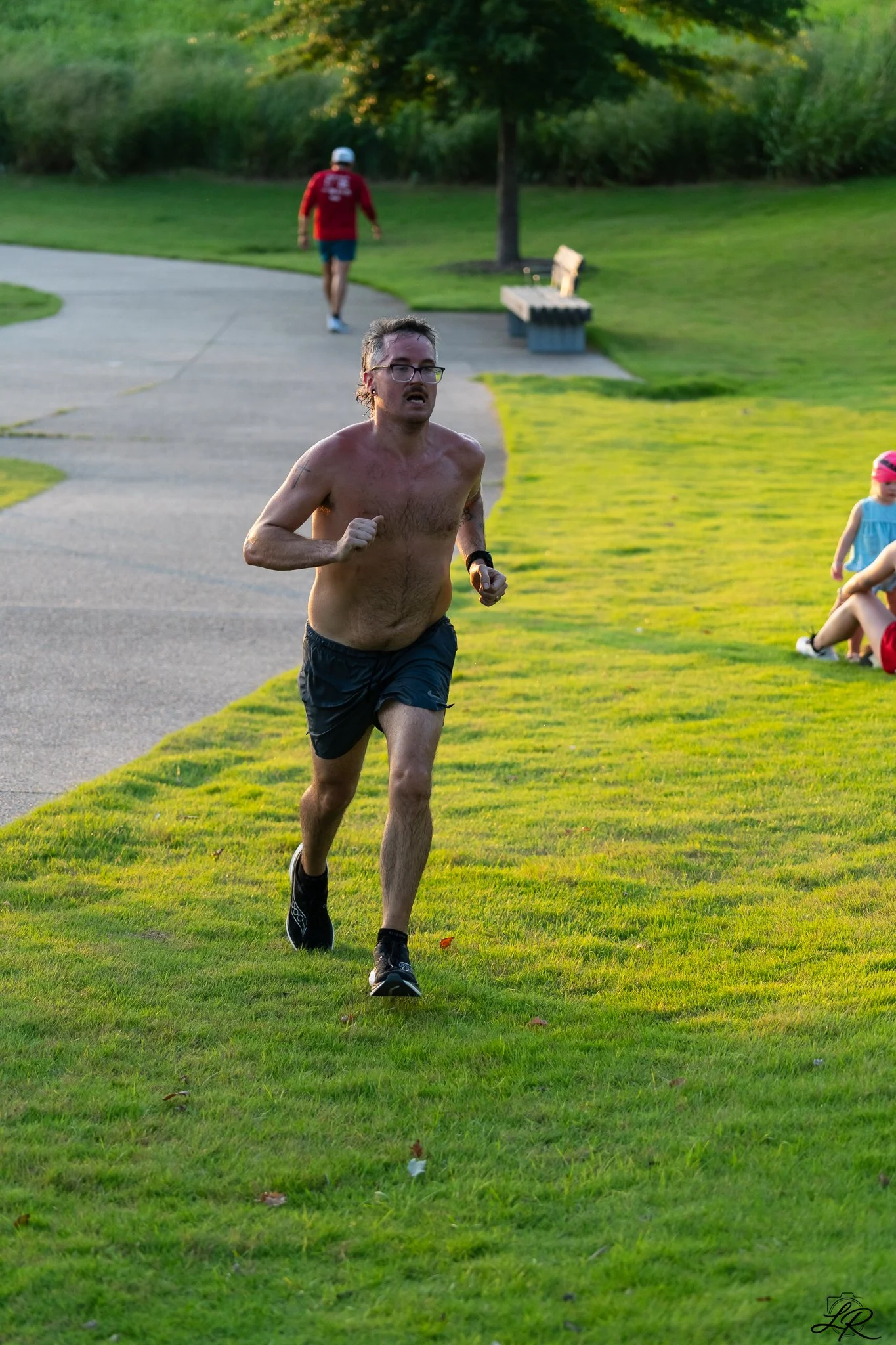 A shirtless man running on a grassy path in a park with trees, with a man in a red shirt and blue shorts walking in the background, and children sitting on the grass to the right.