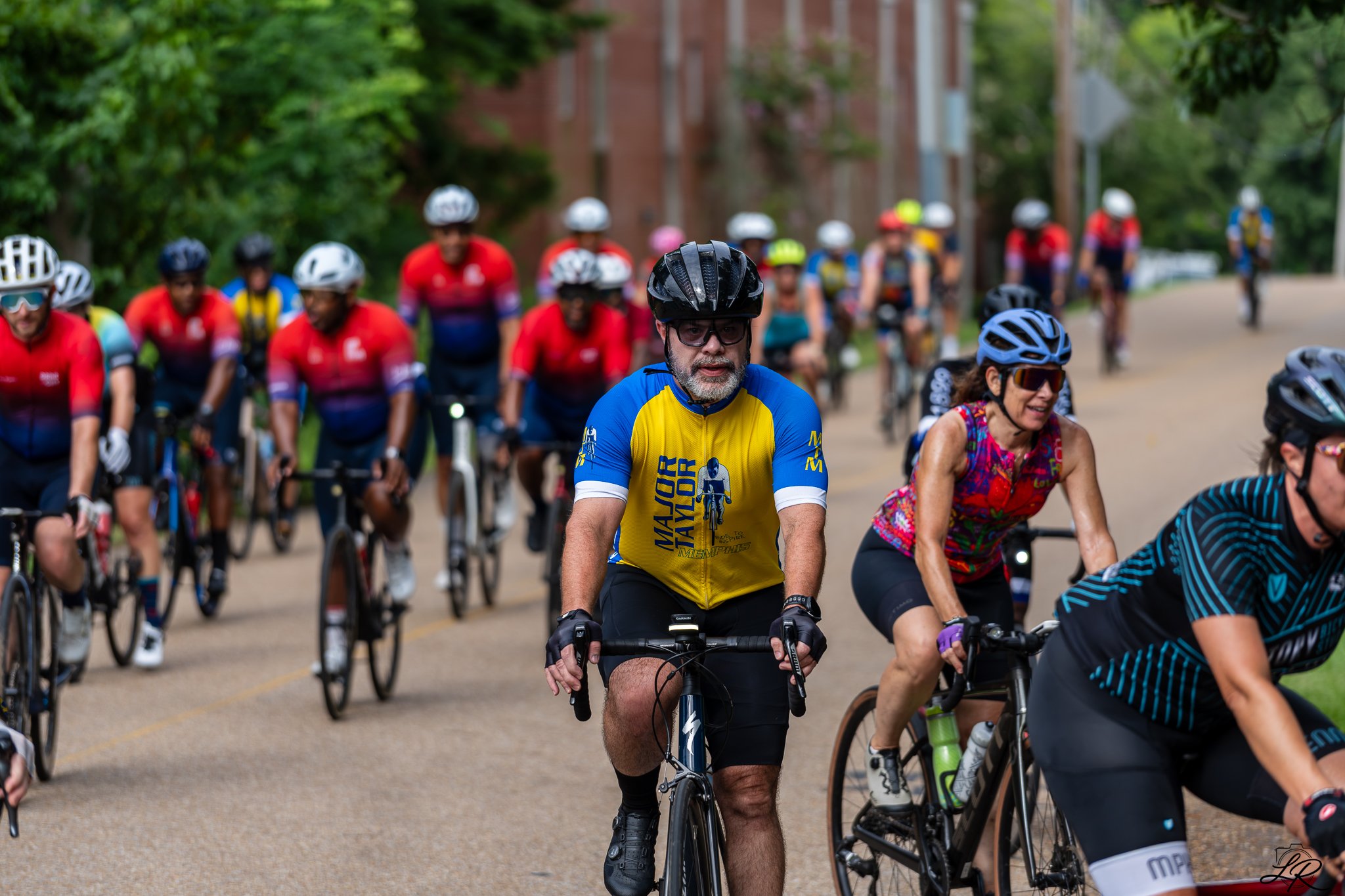 A group of people riding bicycles in a parade or group ride on a street lined with trees and brick buildings.