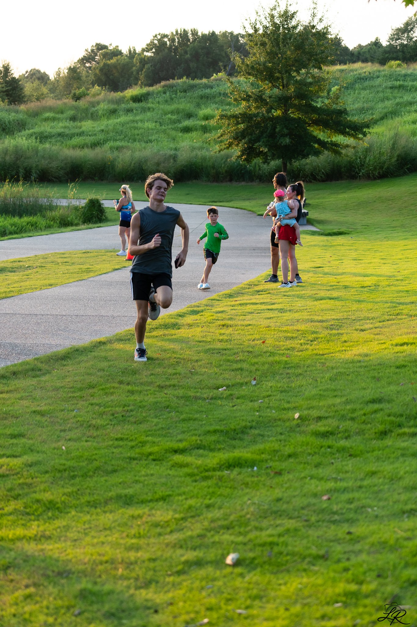 A park with a grassy area, a paved pathway, and people enjoying outdoor activities. One person is running, and others are standing or walking, some with children. There are trees and green hills in the background, illuminated by warm sunlight.