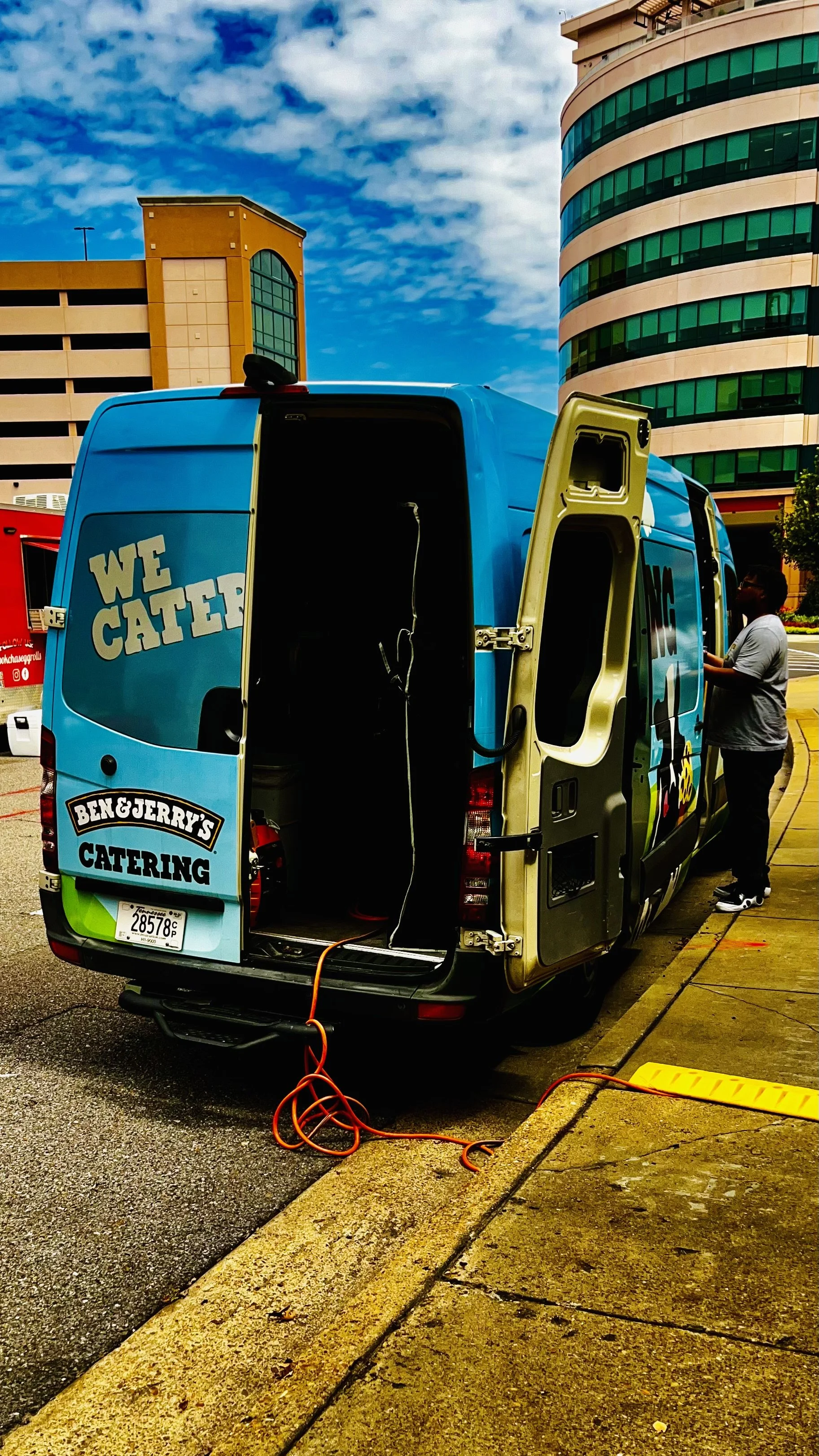 Ben & Jerry's catering van with open side door, parked on a city street near tall buildings, with a person standing next to it and an orange extension cord on the ground.