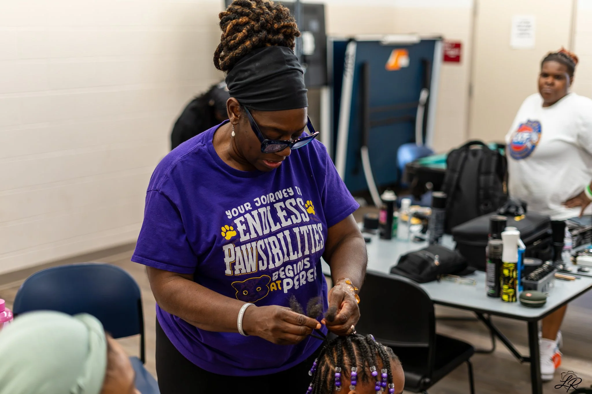 A woman with glasses and curly hair styled in an updo is tying the hair of a child with braided hair with purple beads. The woman is wearing a purple T-shirt that says 'Your journey to endless possibilities begins at Pore' and is participating in a c