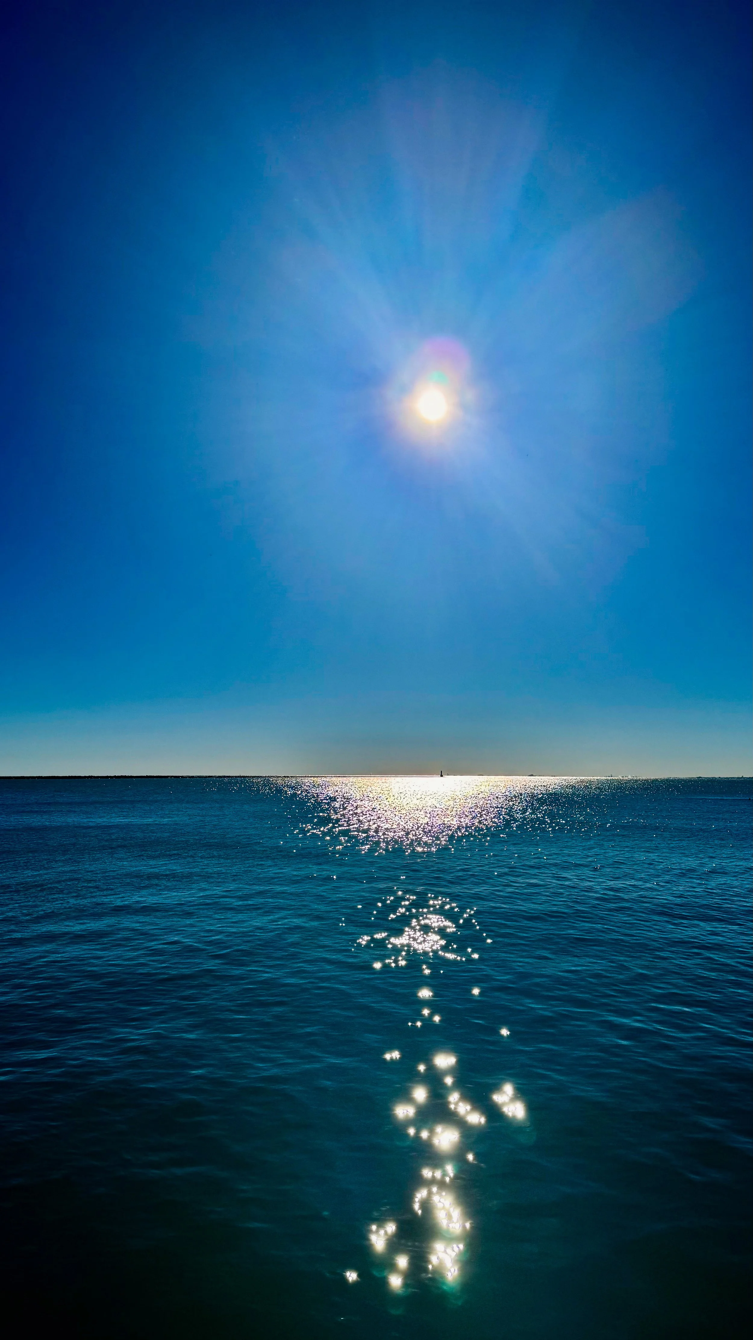 Bright sun shining over calm ocean water, creating a reflection and a starburst effect in a clear blue sky.