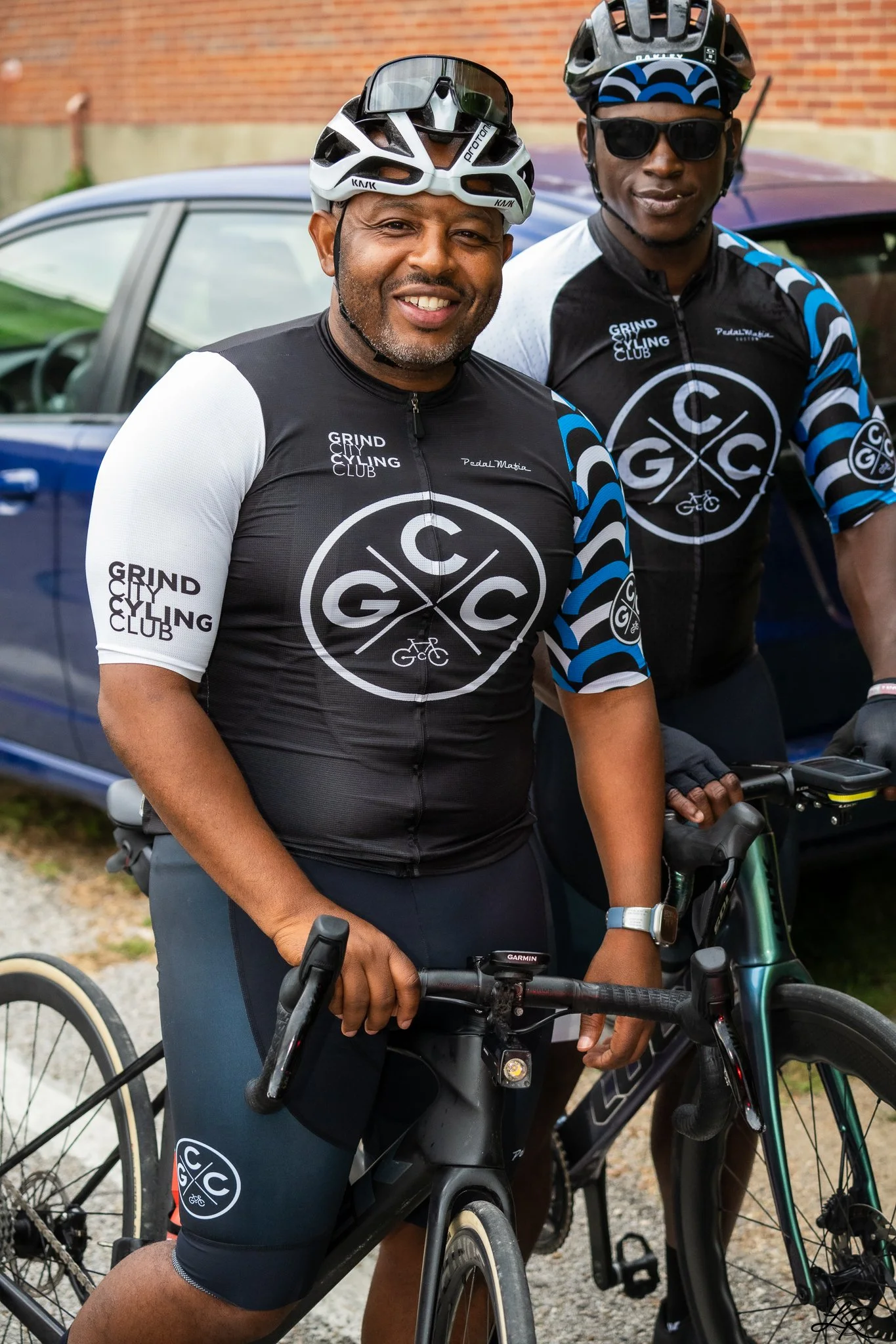 Two men wearing cycling gear and helmets, standing with bikes in front of a car and brick wall. They are smiling and appear ready to ride.