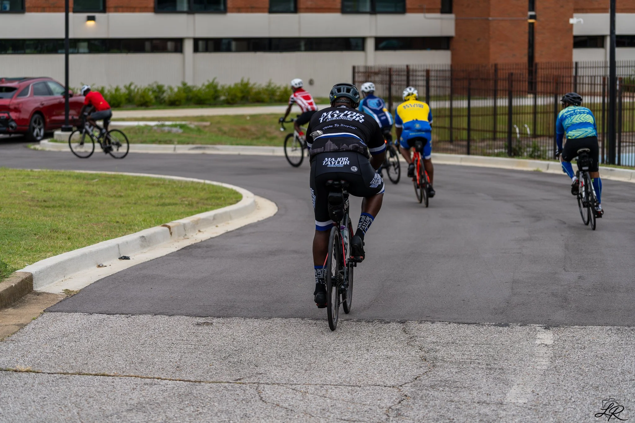 Group of cyclists riding on a curved paved path, wearing colorful jerseys and helmets, with a modern building, parked car, and greenery in the background.
