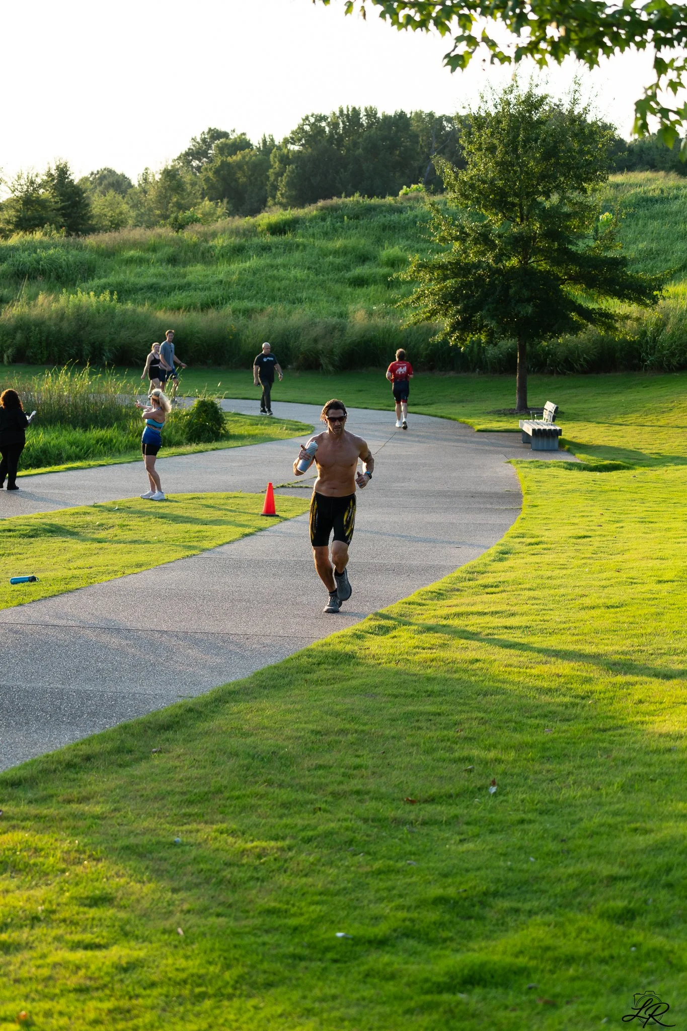 A shirtless man running on a paved path in a park during late afternoon or early evening. Several other people are walking, jogging, or standing along the pathway. The scene is lush with green grass, trees, and a hill in the background, with the sunl