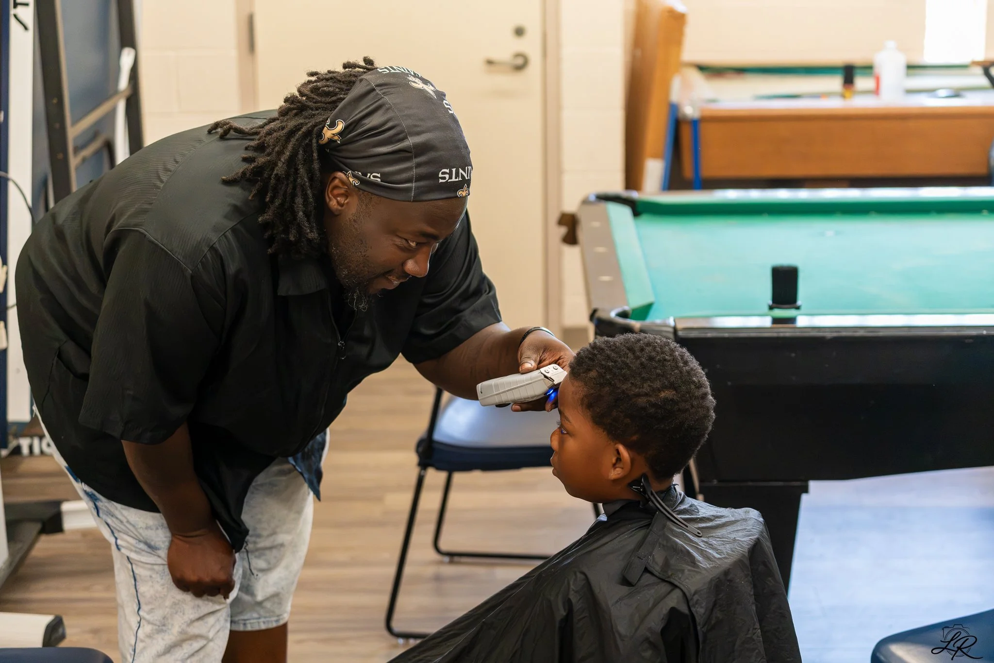 A barber, wearing a black bandana and a black shirt, is leaning over a young boy with curly hair, who is sitting in a barber's chair wearing a black barber cape. The barber is examining or trimming the boy's hair with a measuring tool, inside a barbe
