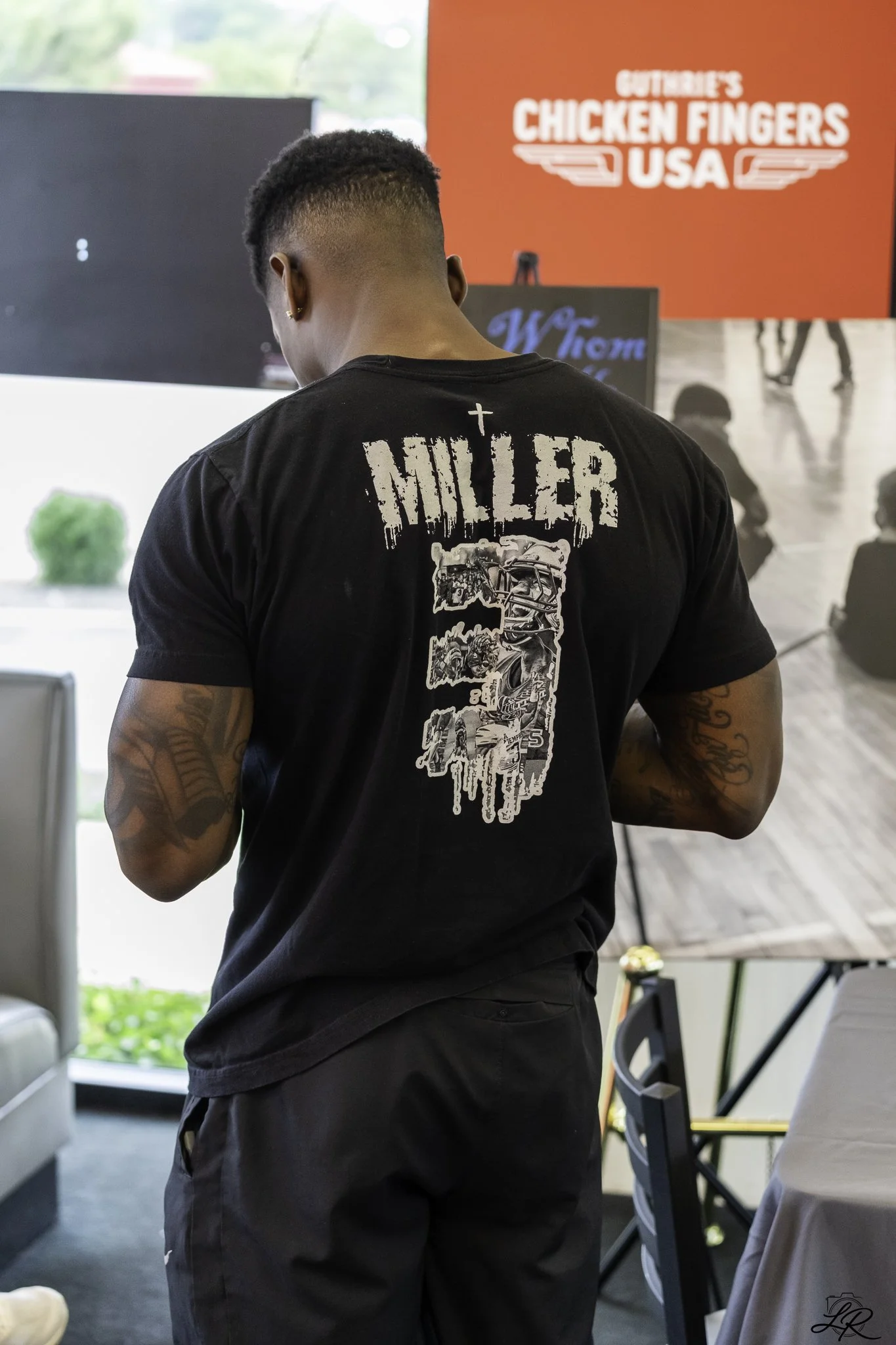 A person with short, black hair and tattoos on their arms, wearing a black t-shirt with the word "MILLER" and a graphic design on the back, standing indoors near a table, with a red sign that says "GUTHRIE'S CHICKEN FINGERS USA" in the background.