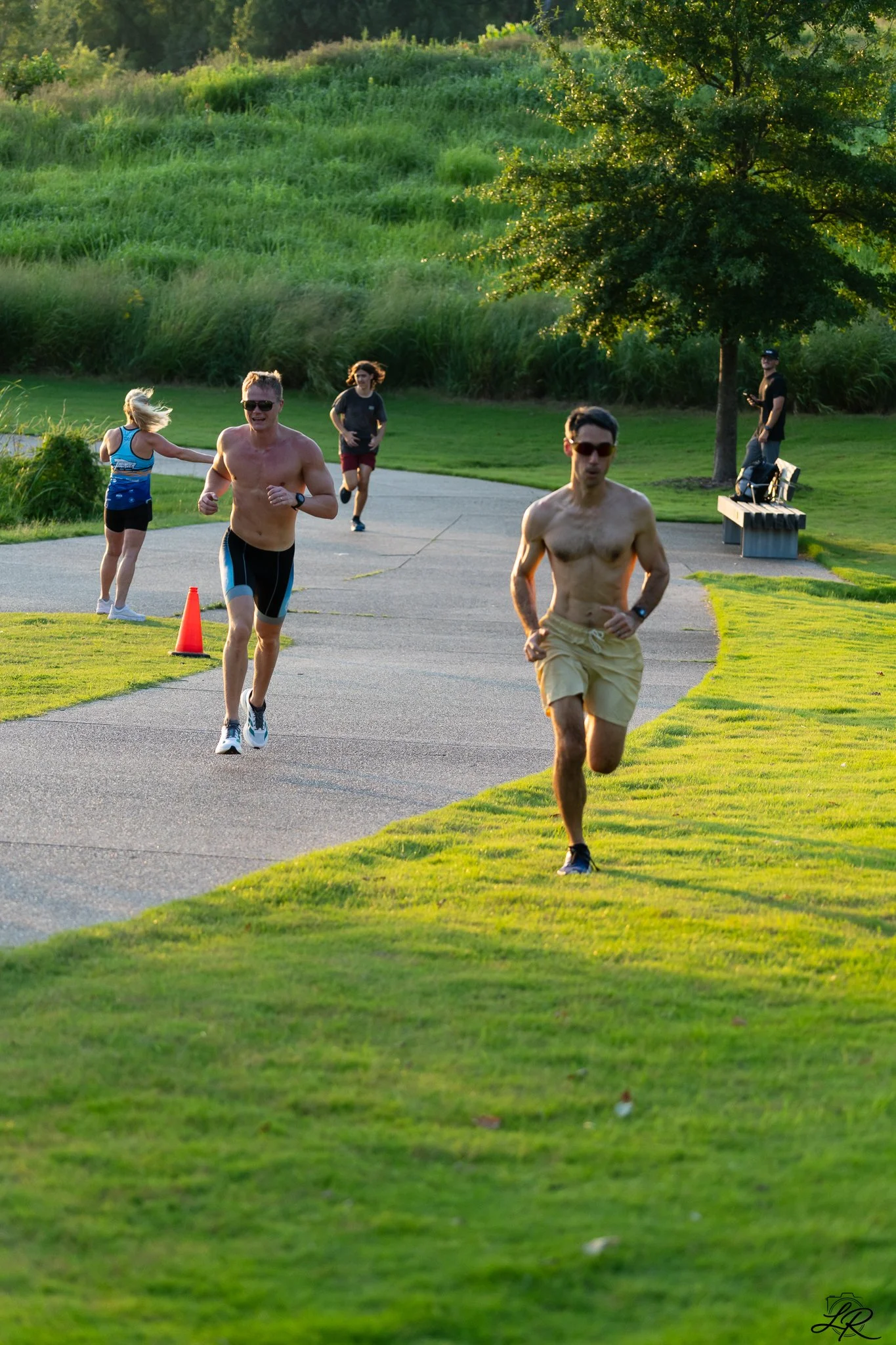 People jogging and running in a park during sunset with green grass, trees, and a bench.
