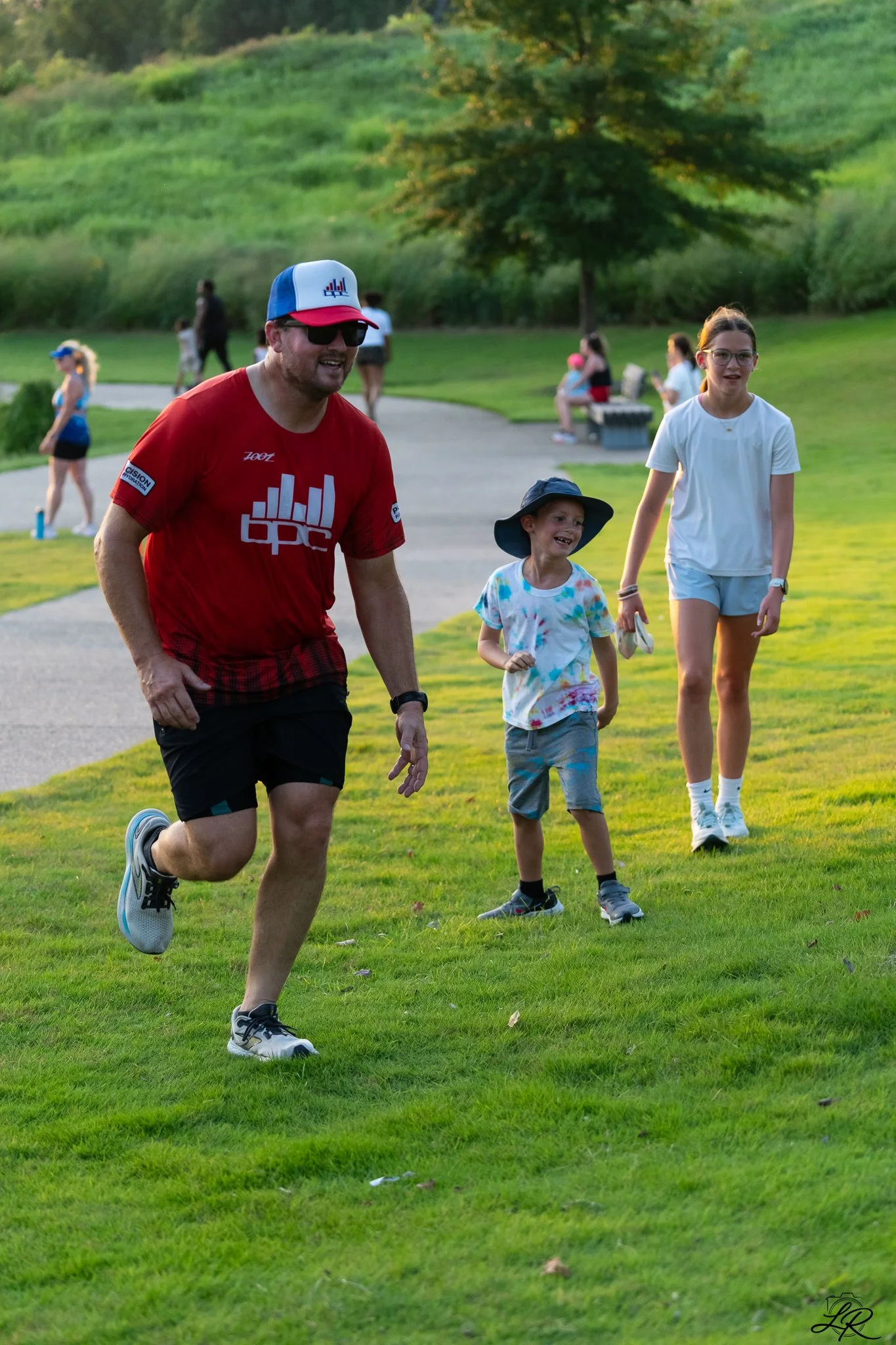 A man running on a grassy area in a park with three young girls walking behind him, during a sunny evening.