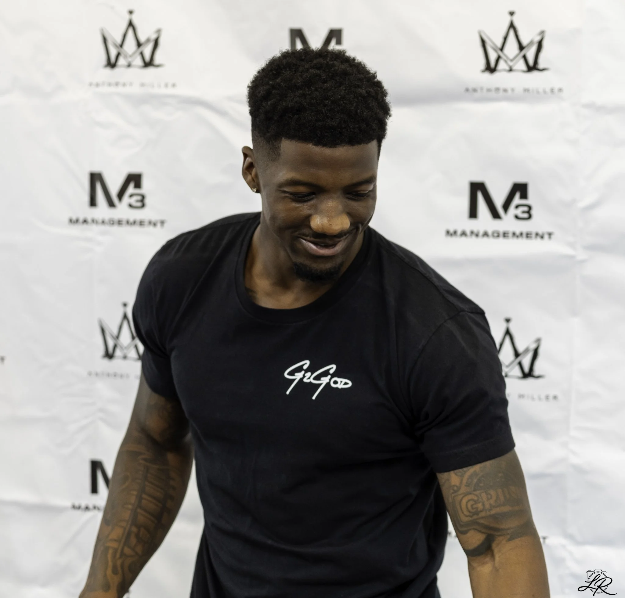 A young man with dark curly hair and tattoos on his arms, smiling while looking down, wearing a black T-shirt with white text, standing in front of a white backdrop with black logos and writing.