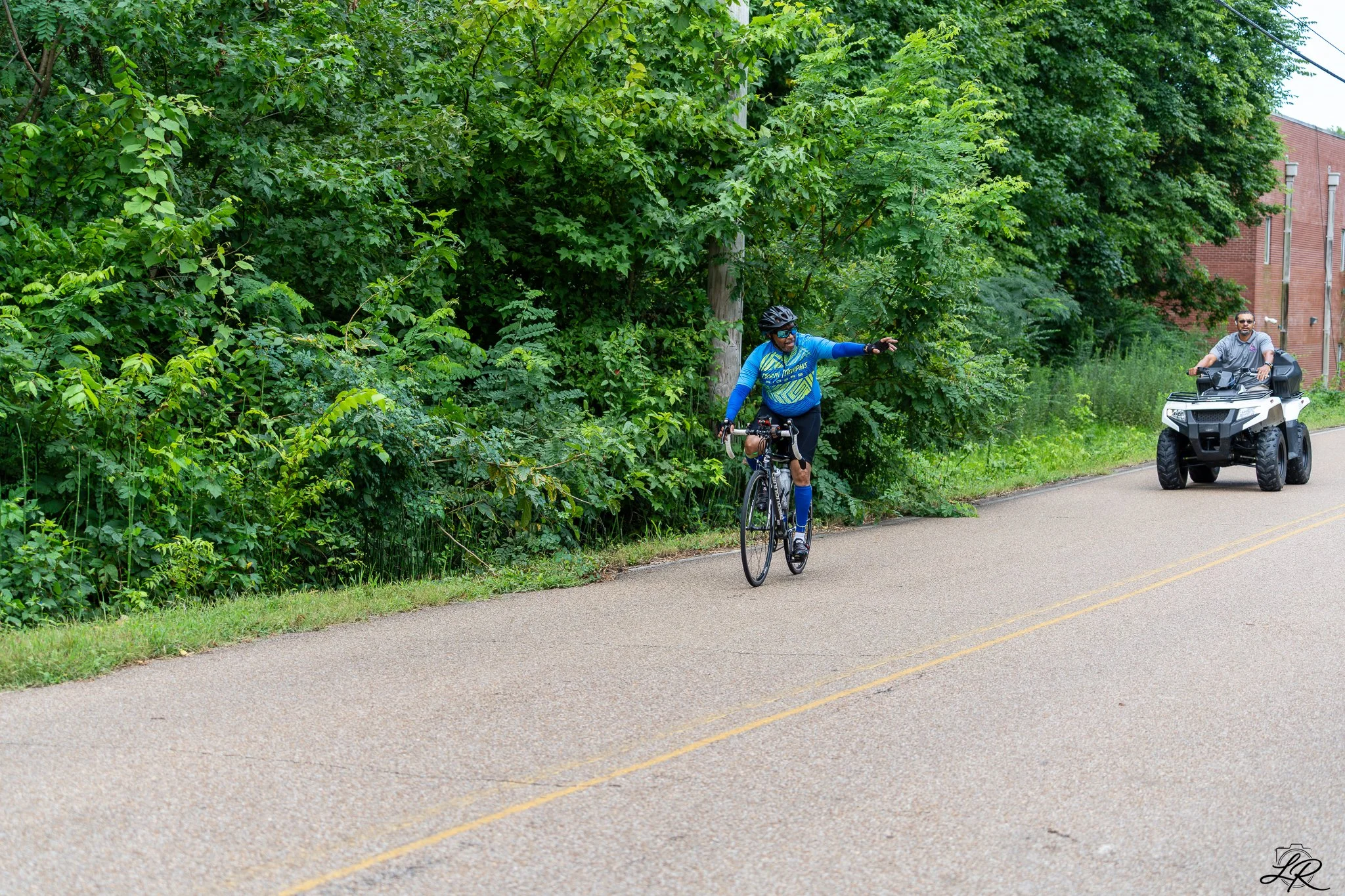A man riding a bicycle and a person on an all-terrain vehicle on a paved road next to a wooded area with green trees and bushes.