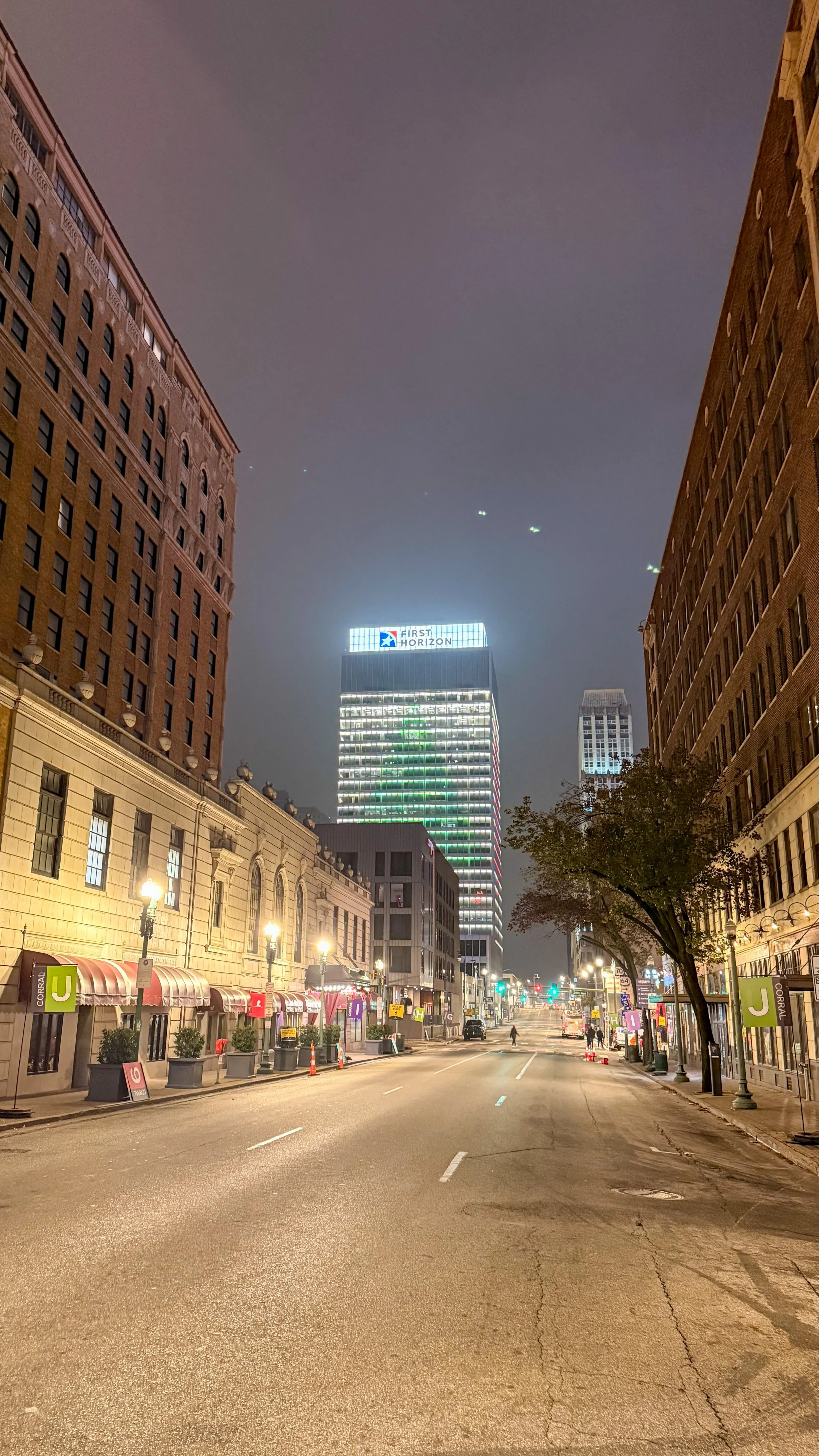 Empty city street at night with buildings lit up, including a tall building with signs for 'First Horizon' and a green-lit hotel. Street lamps illuminate the sidewalk and some trees are visible on the right.