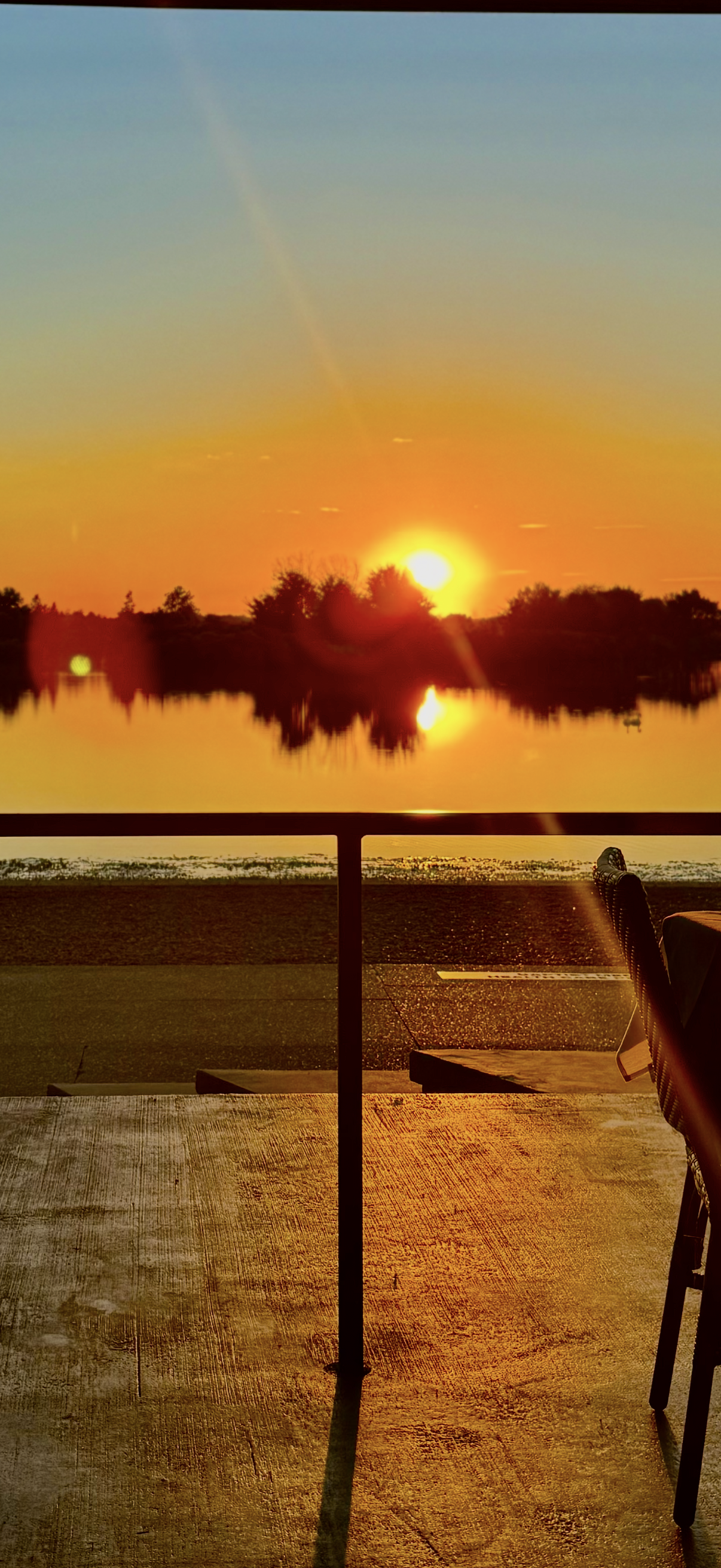 Sunset over a lake, with trees silhouetted along the horizon, reflected in the water, viewed from a patio with a railing and part of a chair.