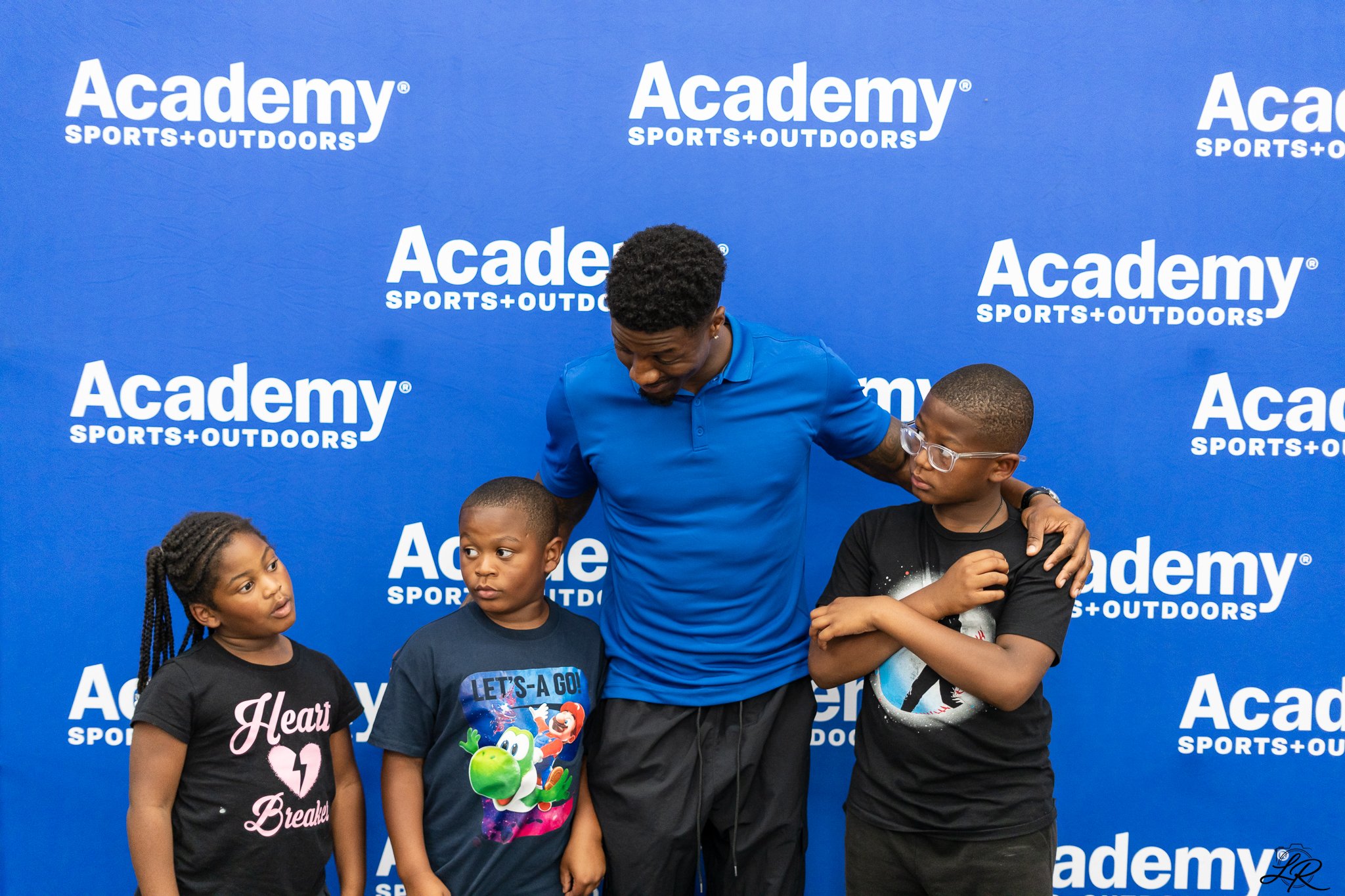 A man in a blue shirt smiling and interacting with three children in front of an Academy Sports + Outdoors backdrop. The children include a girl with braided hair and a shirt that says 'Heart Breaker,' a boy wearing a 'Let's a Go!' shirt featuring Ma
