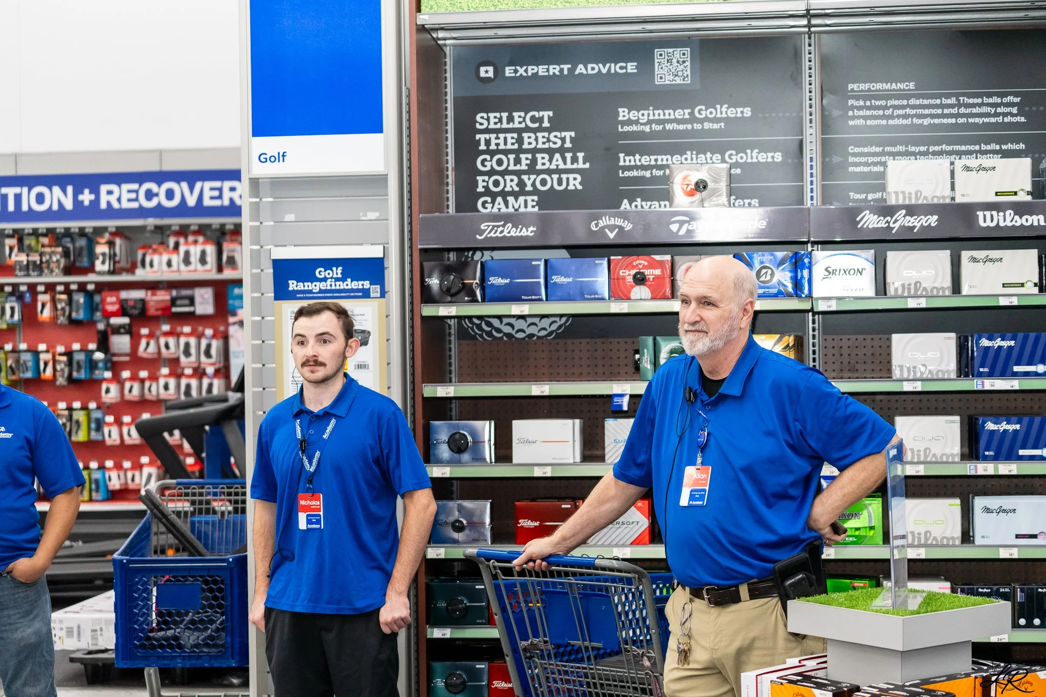 Two male employees in blue polo shirts working at a golf equipment section in a retail store, surrounded by golf balls, golf clubs, and related accessories on shelves.
