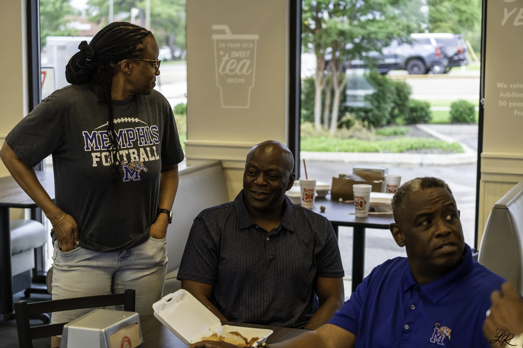 A woman standing and smiling while talking to two seated men at a table inside a fast food restaurant. The woman is wearing a Memphis Football t-shirt, glasses, and has braided hair. The man in the center has a bald head and is smiling, wearing a bla