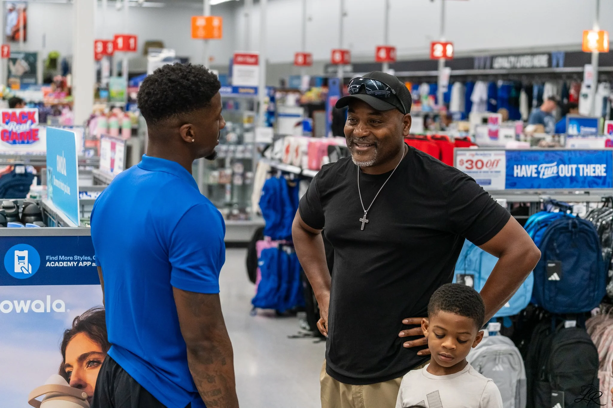 Two men and two children inside a retail store, engaged in conversation. The man on the left in a blue shirt is facing the man on the right, who is wearing a black shirt, a black cap, sunglasses, and a cross necklace. The children appear to be listen