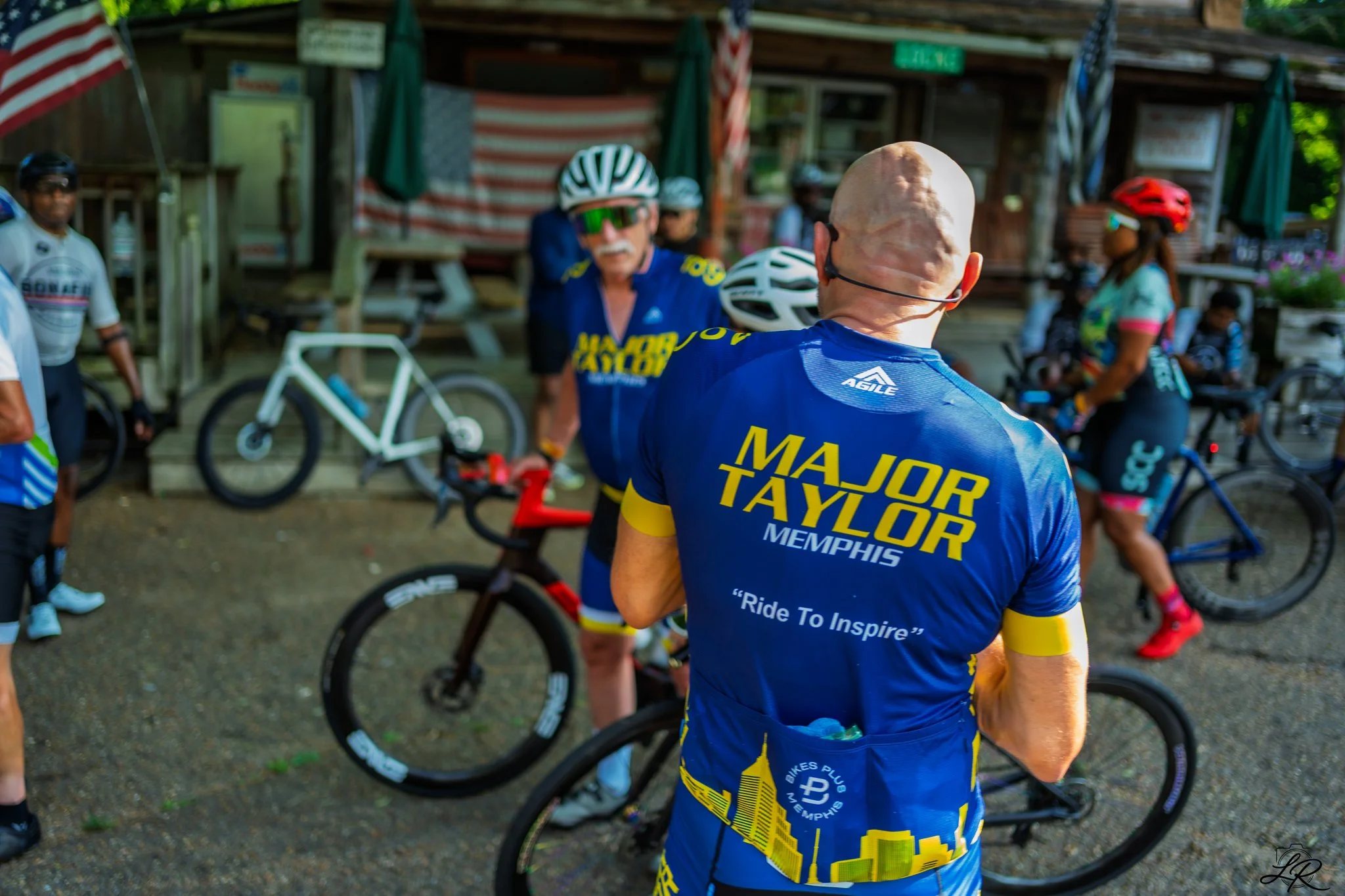 Cyclists gathered outdoors, with one cyclist in the foreground wearing a blue and yellow jersey reading 'Major Taylor Memphis' and a helmet, surrounded by others with bicycles and helmets.