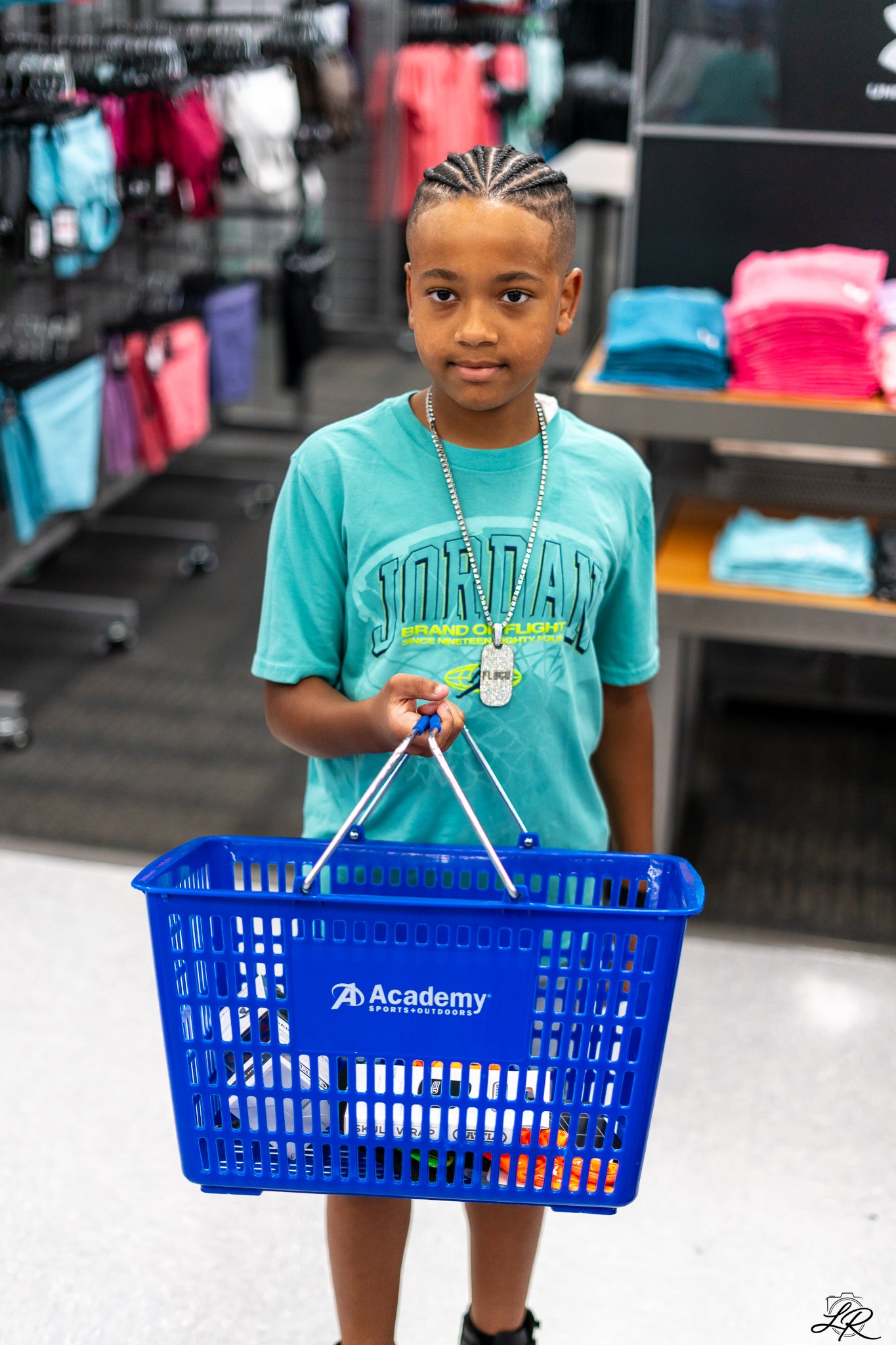 A young boy with cornrow braids and a turquoise T-shirt holding a blue shopping basket at Academy Sports + Outdoors store.