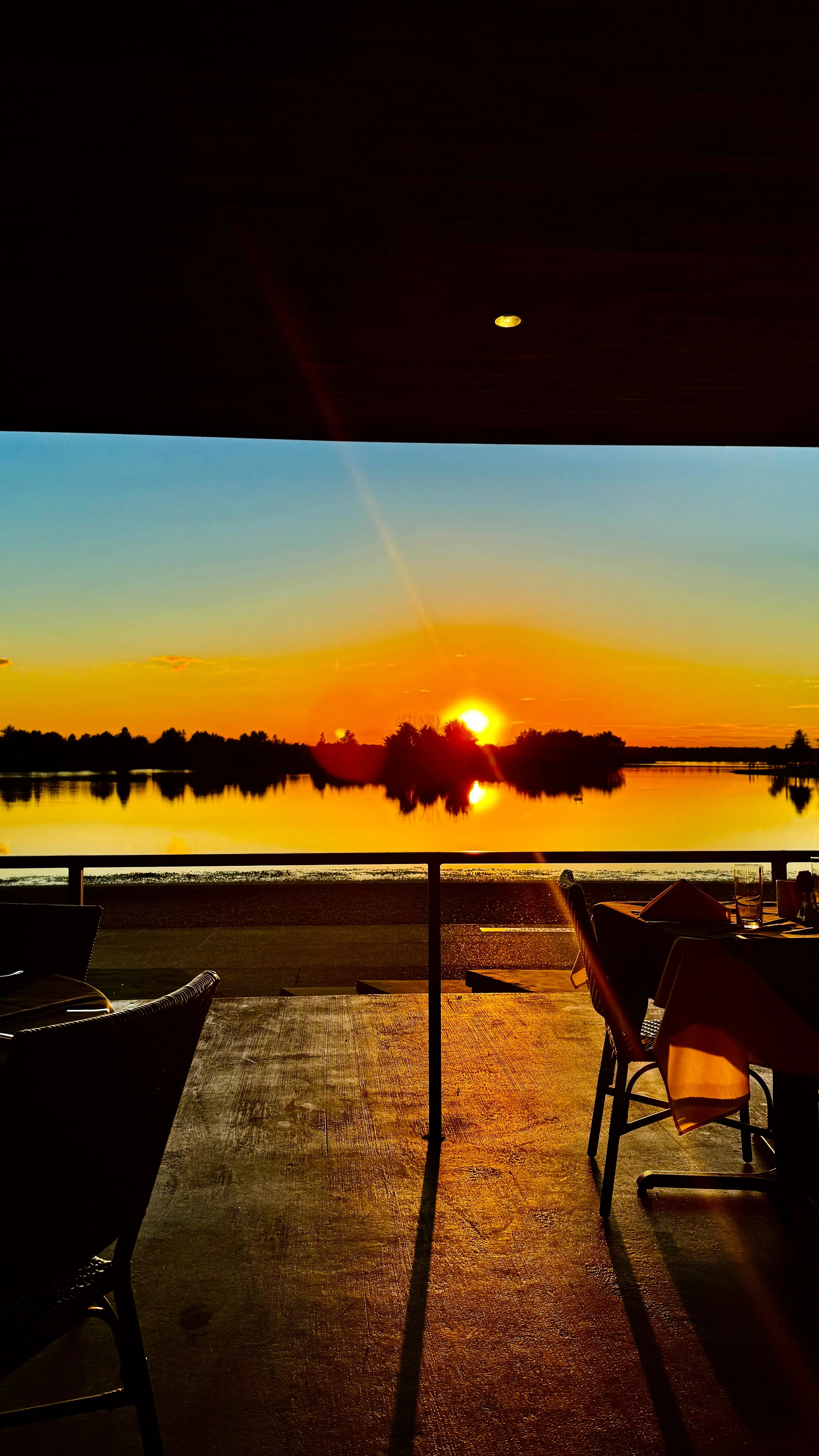 A scenic sunset over a calm body of water with silhouetted trees, viewed from a restaurant patio with tables and chairs.
