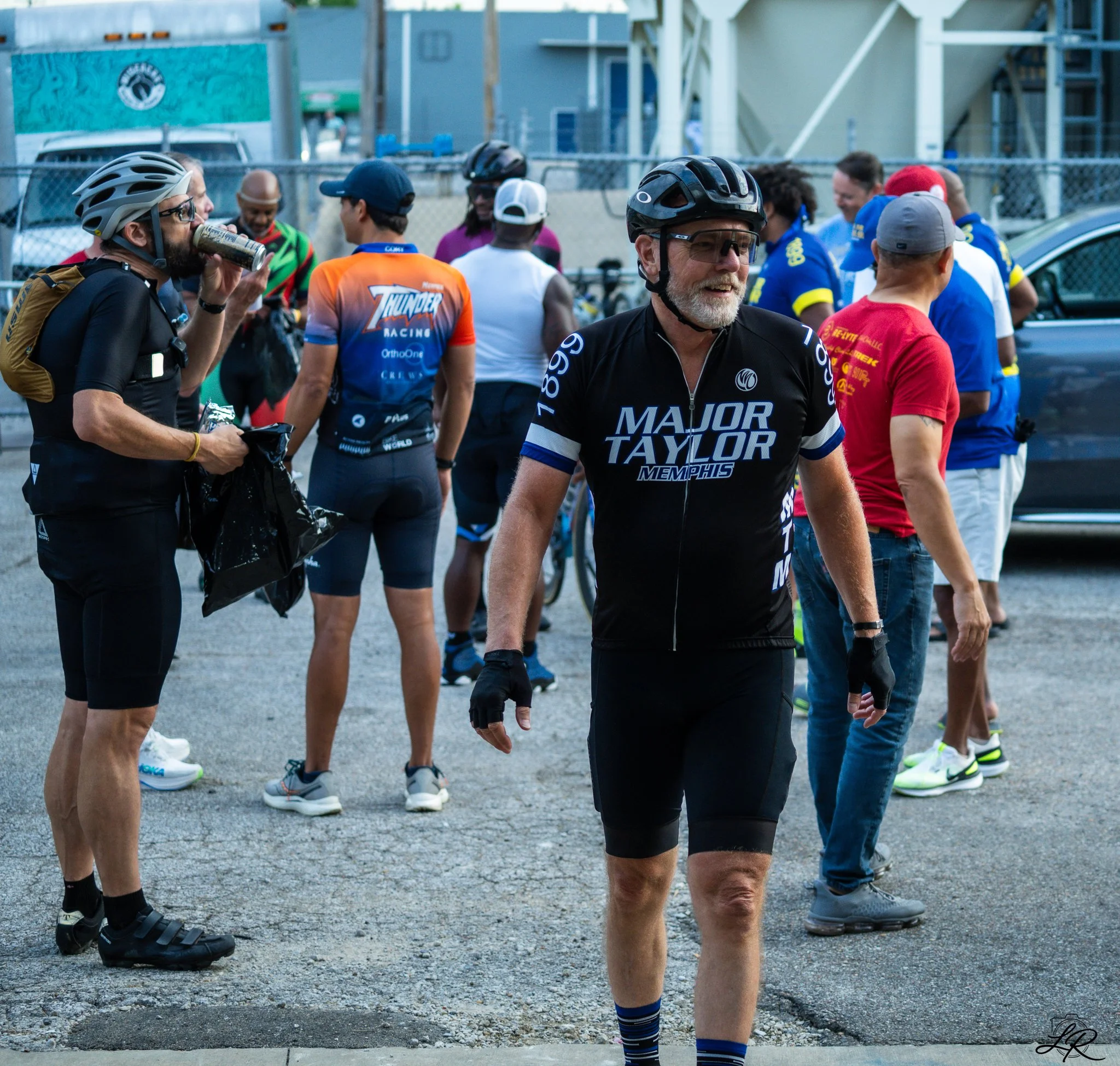 Group of people wearing cycling gear and helmets, standing on a gravel area outdoors.