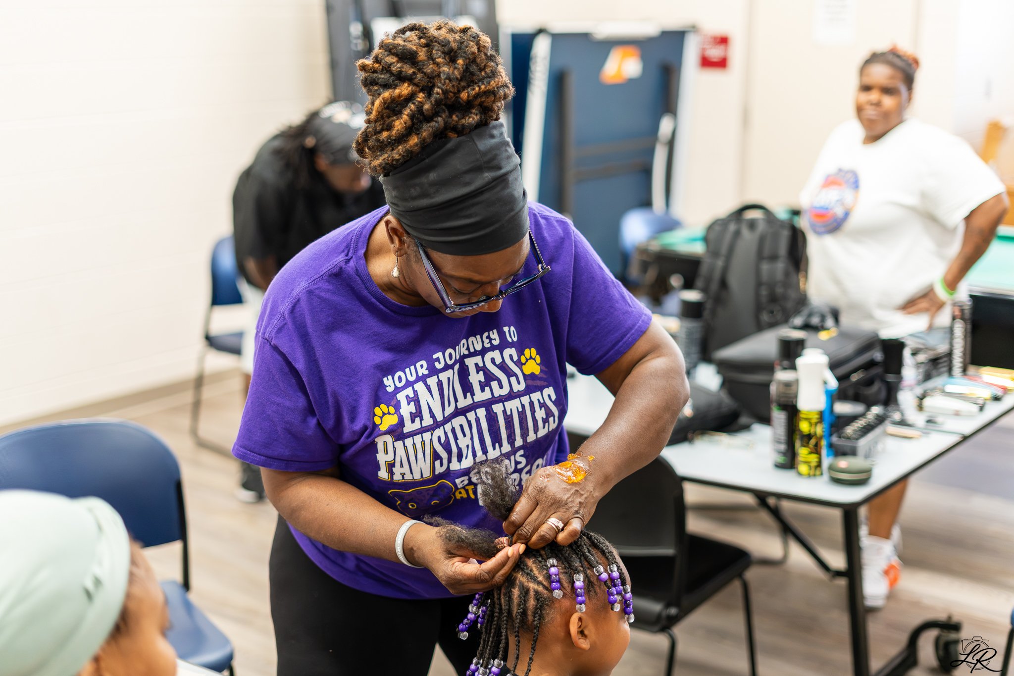 A woman with a head wrap and glasses braiding or styling a child's dreadlocks with beads in a classroom or community center setting, with other women and supplies in the background.