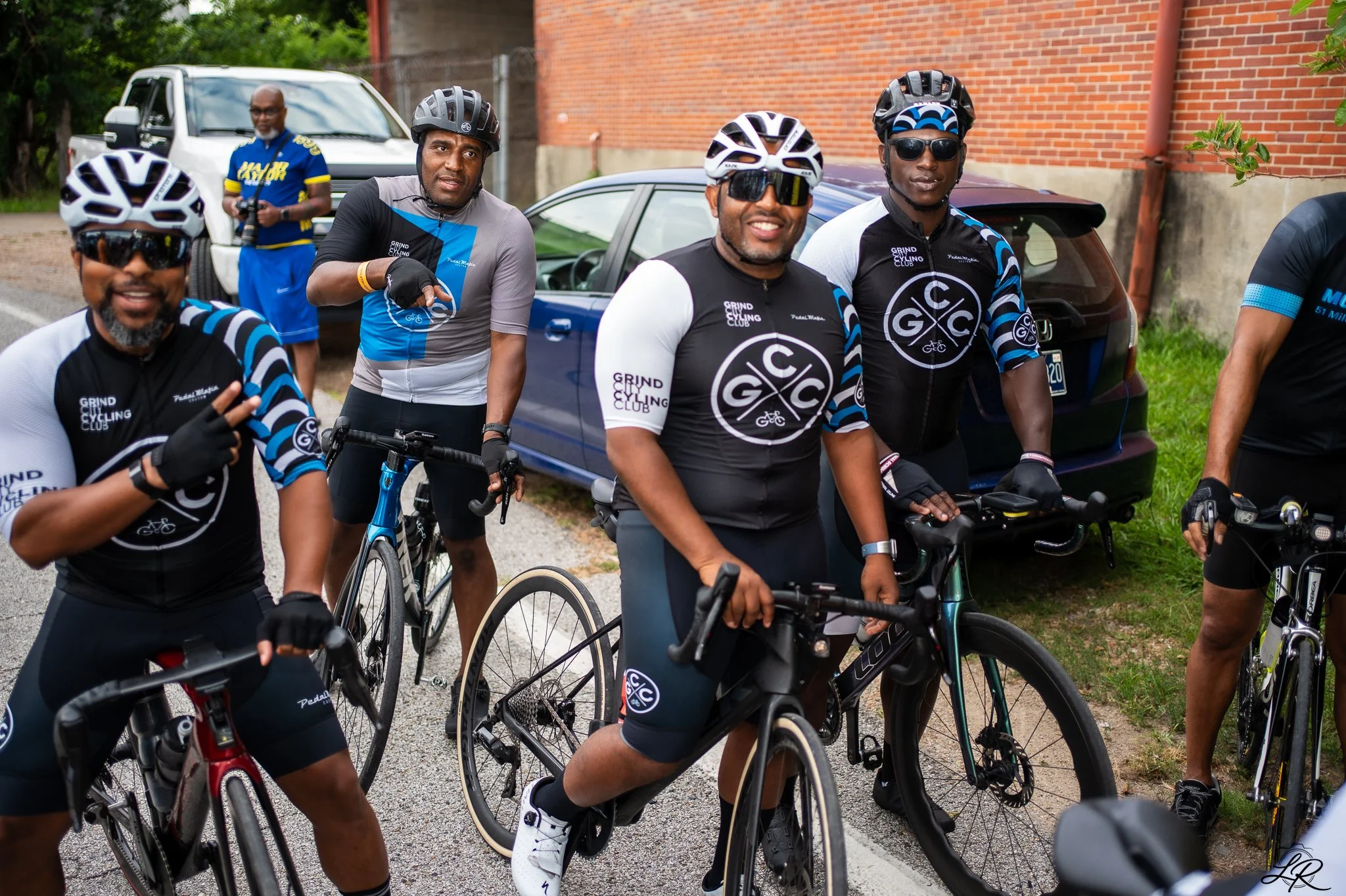 A group of cyclists wearing helmets and sunglasses, standing with their bikes outdoors near a brick wall and parked cars, smiling and looking at the camera.