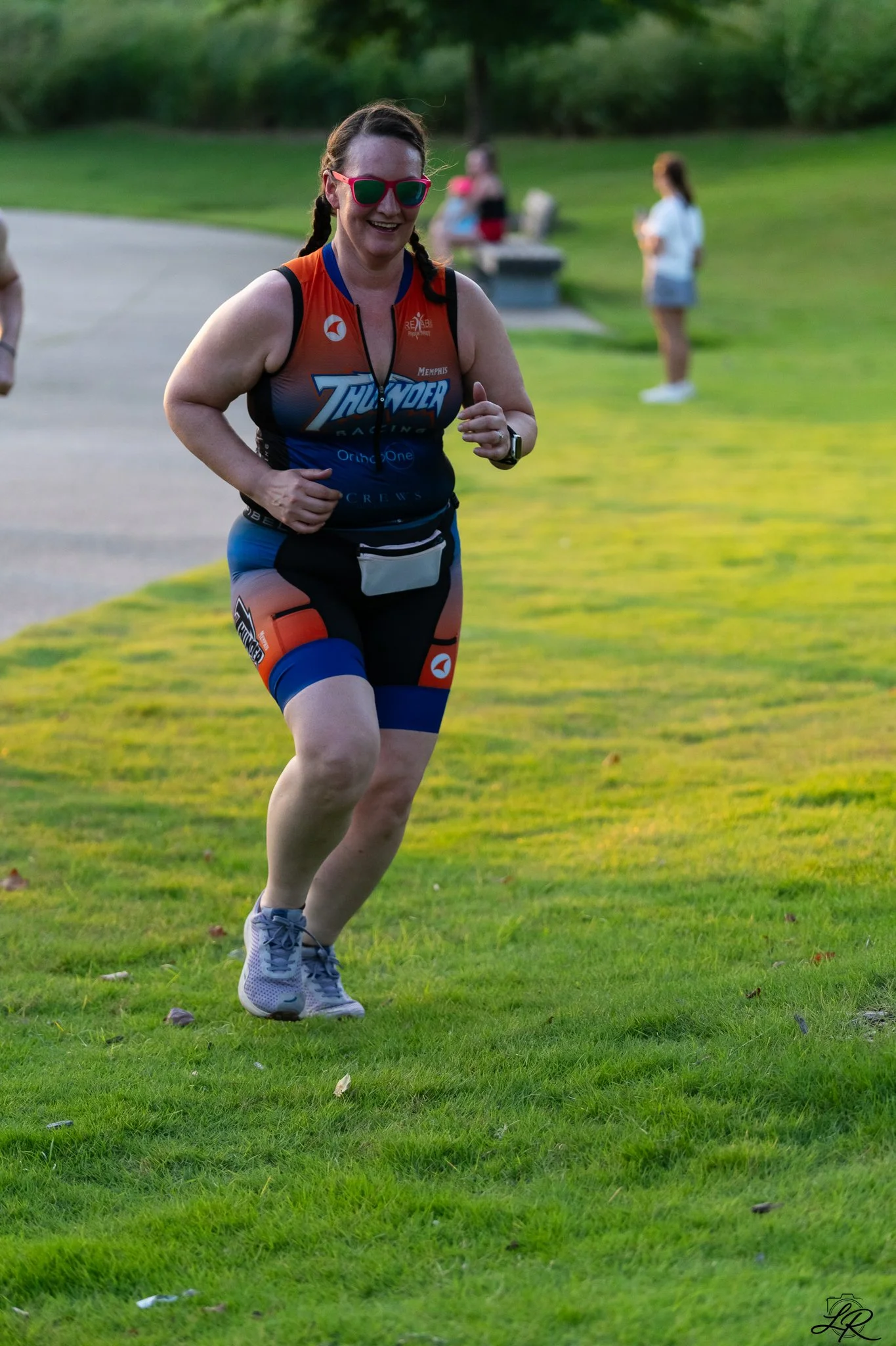 A woman running outdoors on a grassy area, wearing a racing suit with the words "Thunder Racing" and sunglasses, sporting a digital watch, with two women in the background standing on the grass, one in a white shirt and shorts