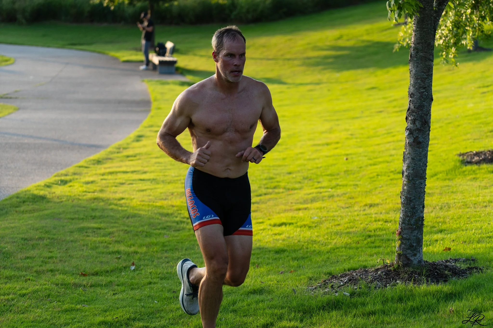 A shirtless man running outdoors on a grassy area with trees, wearing running shorts and shoes, with a person in the background near a park bench.