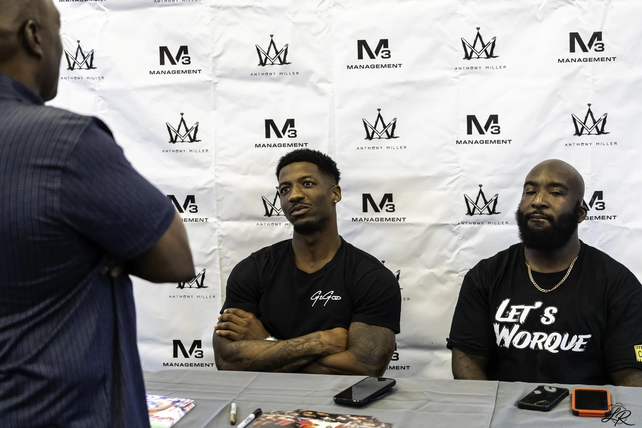 Two men sitting at a table with a third person standing in front of them. The backdrop behind them features logos and text including 'Anthony Miller', 'M3 Management', and a crown emblem. The man on the left has his arms crossed, has tattoos on his f