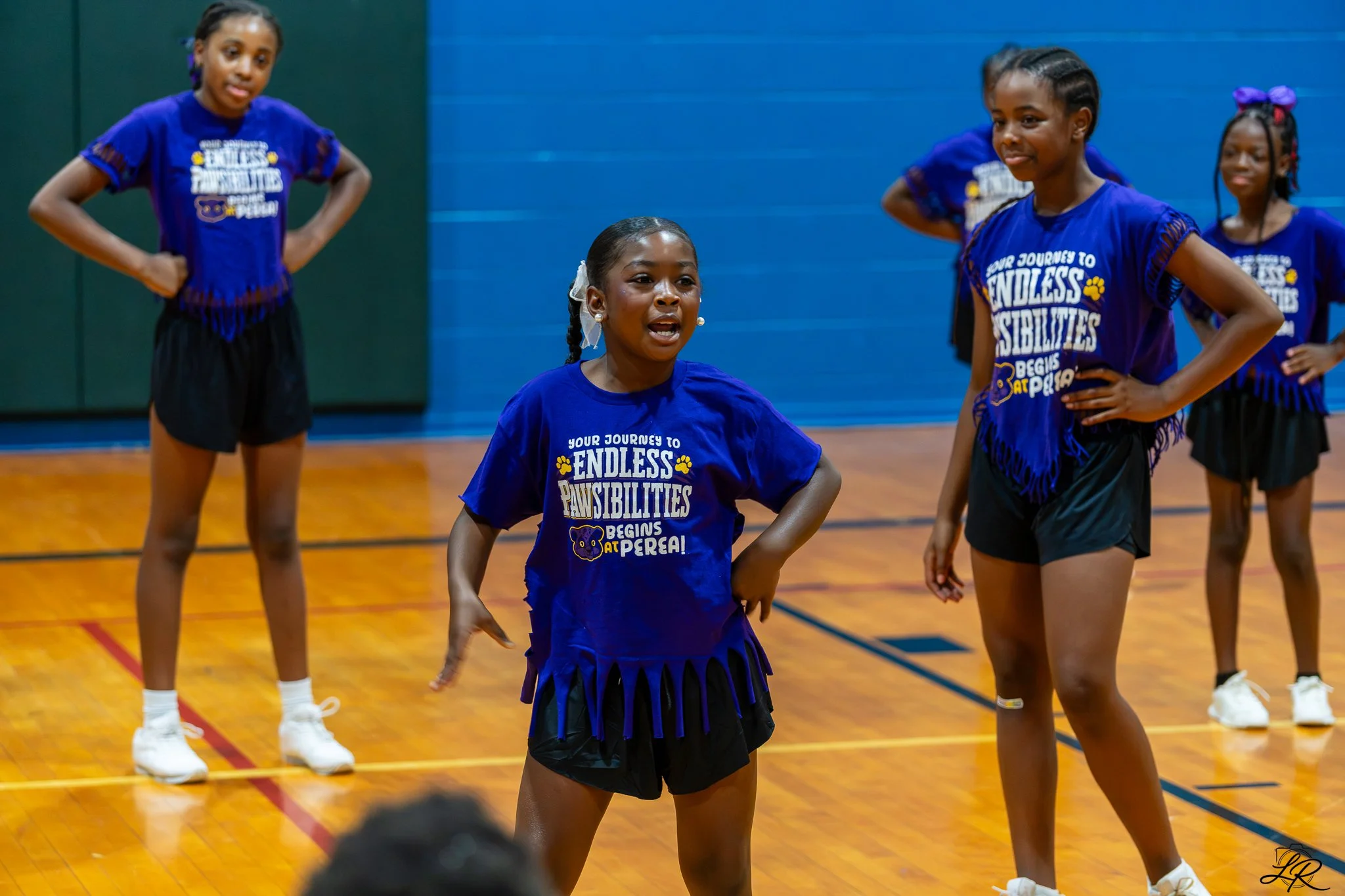 Young girls participating in a cheerleading event in a gymnasium, wearing matching blue t-shirts with the phrase "Your journeys to endless responsibilities begins at Perea" and black skirts. They are standing on a wooden gym floor with colorful lines