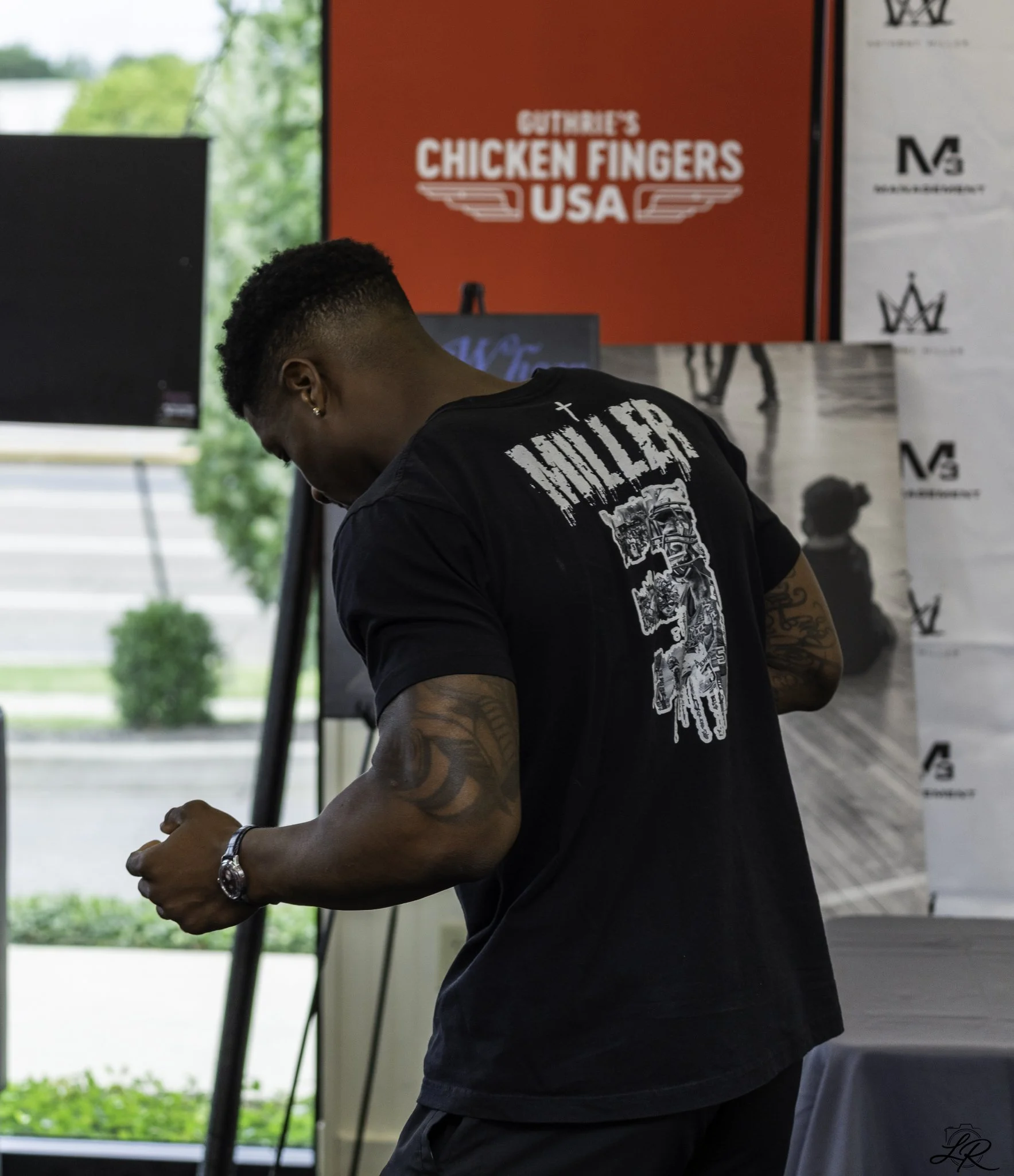 A man wearing a black t-shirt with a graphic design and a wristwatch, standing indoors near tables and banners. The background features a red poster for 'CITRIBE'S CHICKEN FINGERS USA' and other promotional logos, with large windows showing greenery 