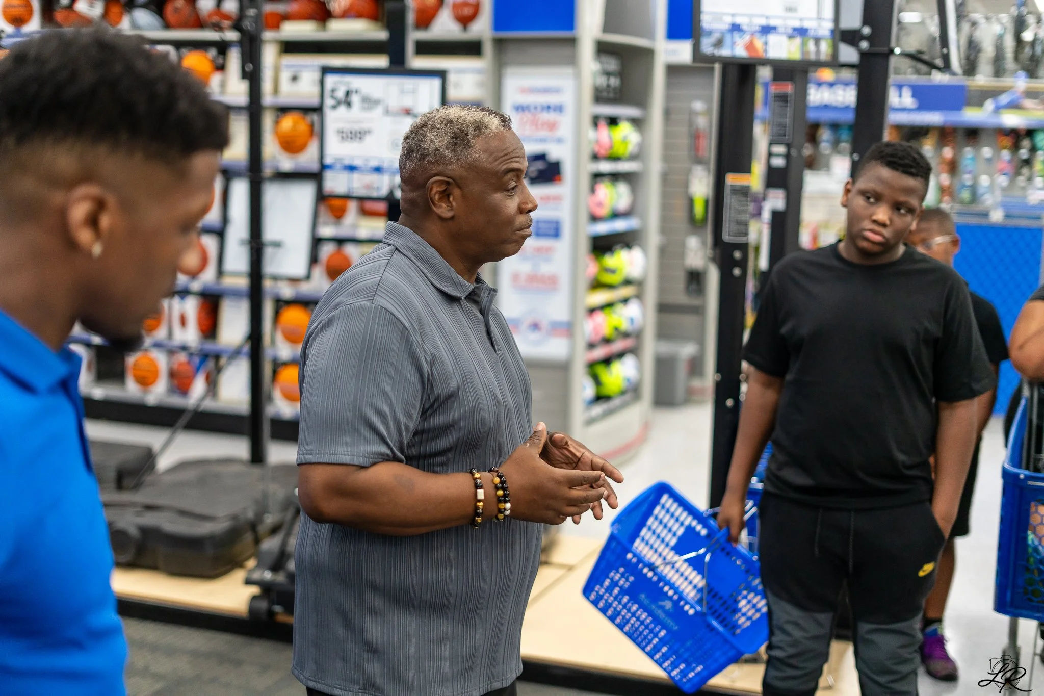 A man with gray hair, wearing a gray striped shirt, standing in a store, engaged in conversation with a group of teenagers. The store has shelves stocked with sports equipment, including basketballs and helmets, and computer monitors. The teenagers a