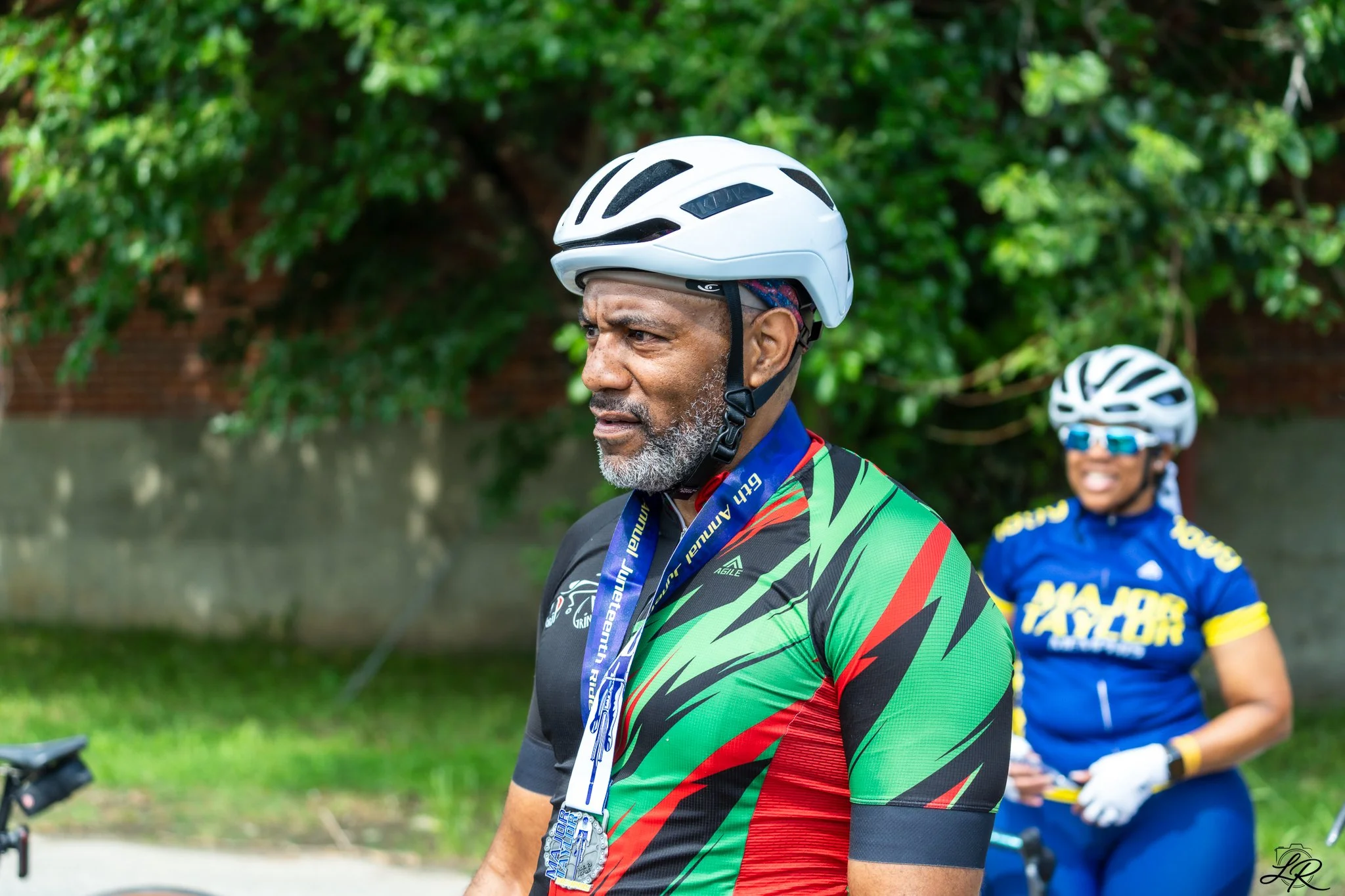 A man with a gray beard wearing a white bike helmet and colorful cycling jersey, outdoors with green foliage in the background.
