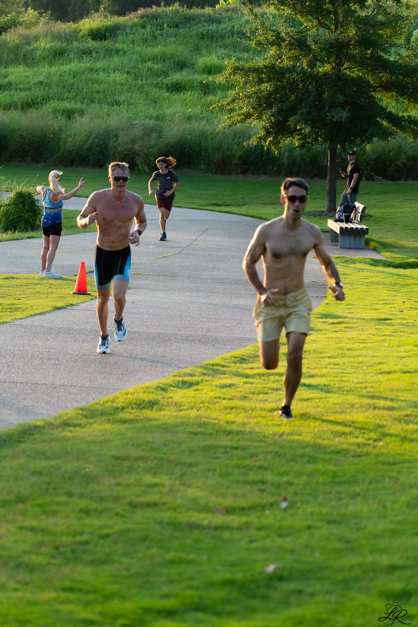 Two men are running in a park on a paved trail, one shirtless and wearing sunglasses, and the other wearing sunglasses and shorts. A woman in a blue top and black shorts is pointing and cheering near a cone on the grass. There are two other people in