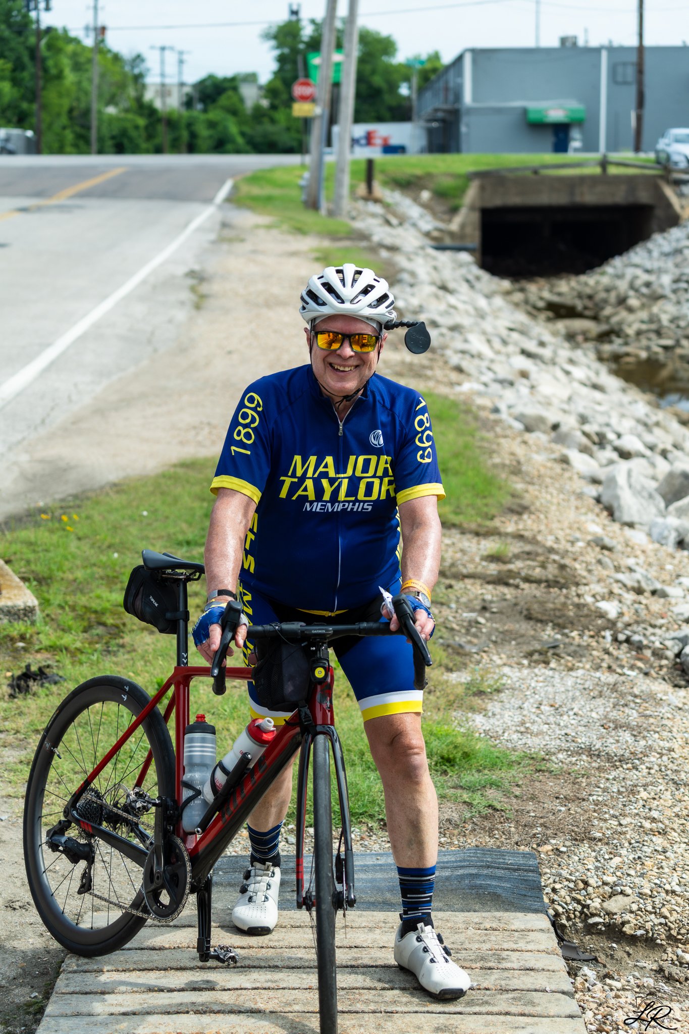A smiling man in cycling gear, wearing a blue and yellow team jersey, white helmet, and sunglasses, standing with a red mountain bike on a scenic outdoor trail near a stream and bridge.