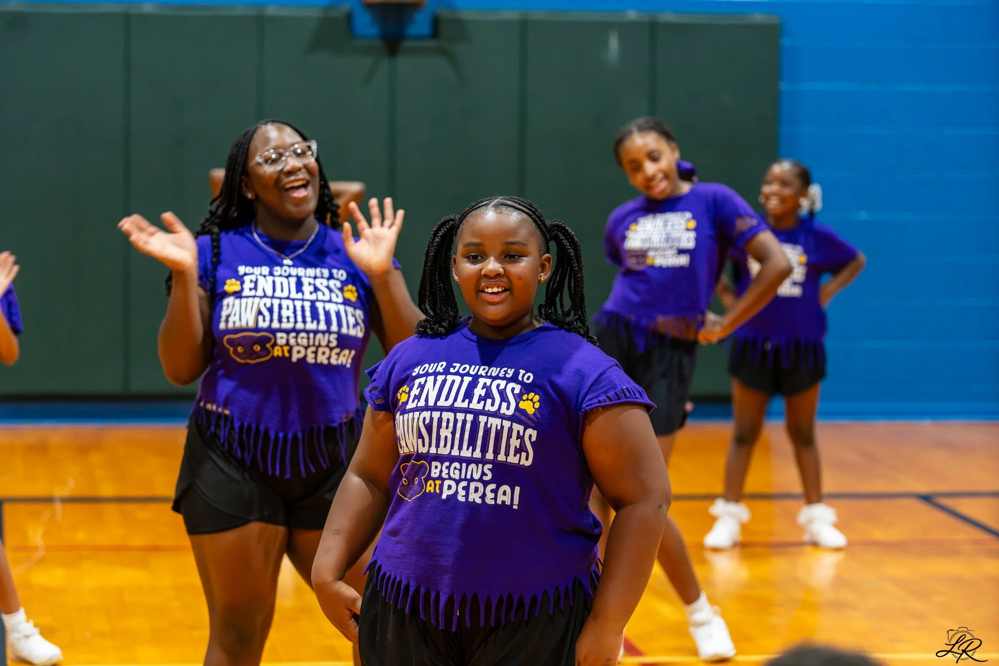 Group of young girls in purple T-shirts dancing and smiling in a gymnasium.